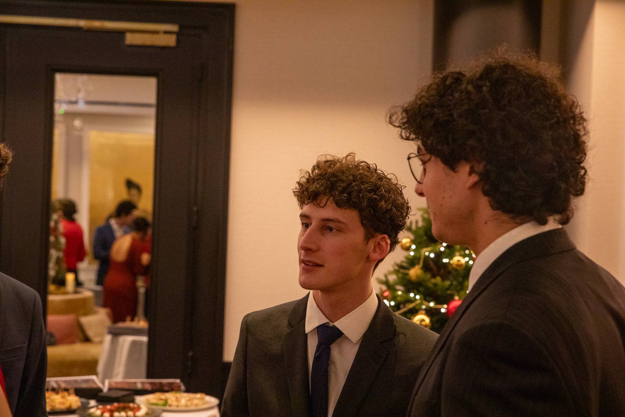 Deux étudiants en costume discutent près d'un buffet, devant un sapin de Noël et un salon éclairé au fond.