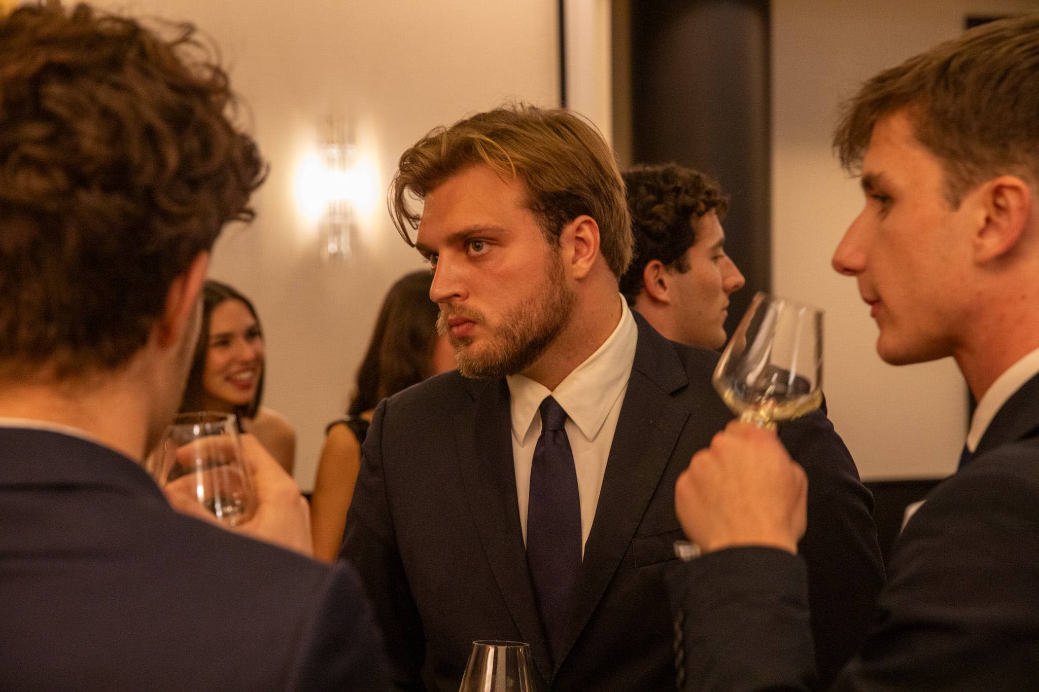 Un groupe d'étudiants en costume conversent debout, verre de vin blanc à la main, dans un salon lambrissé.
