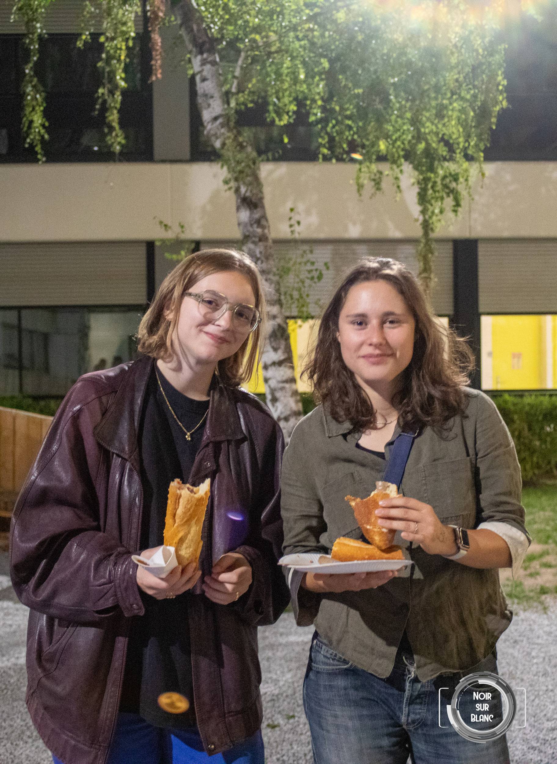 Deux étudiantes debout dans la nuit, une assiette et un sandwich à la main, devant un bâtiment vitré du campus.