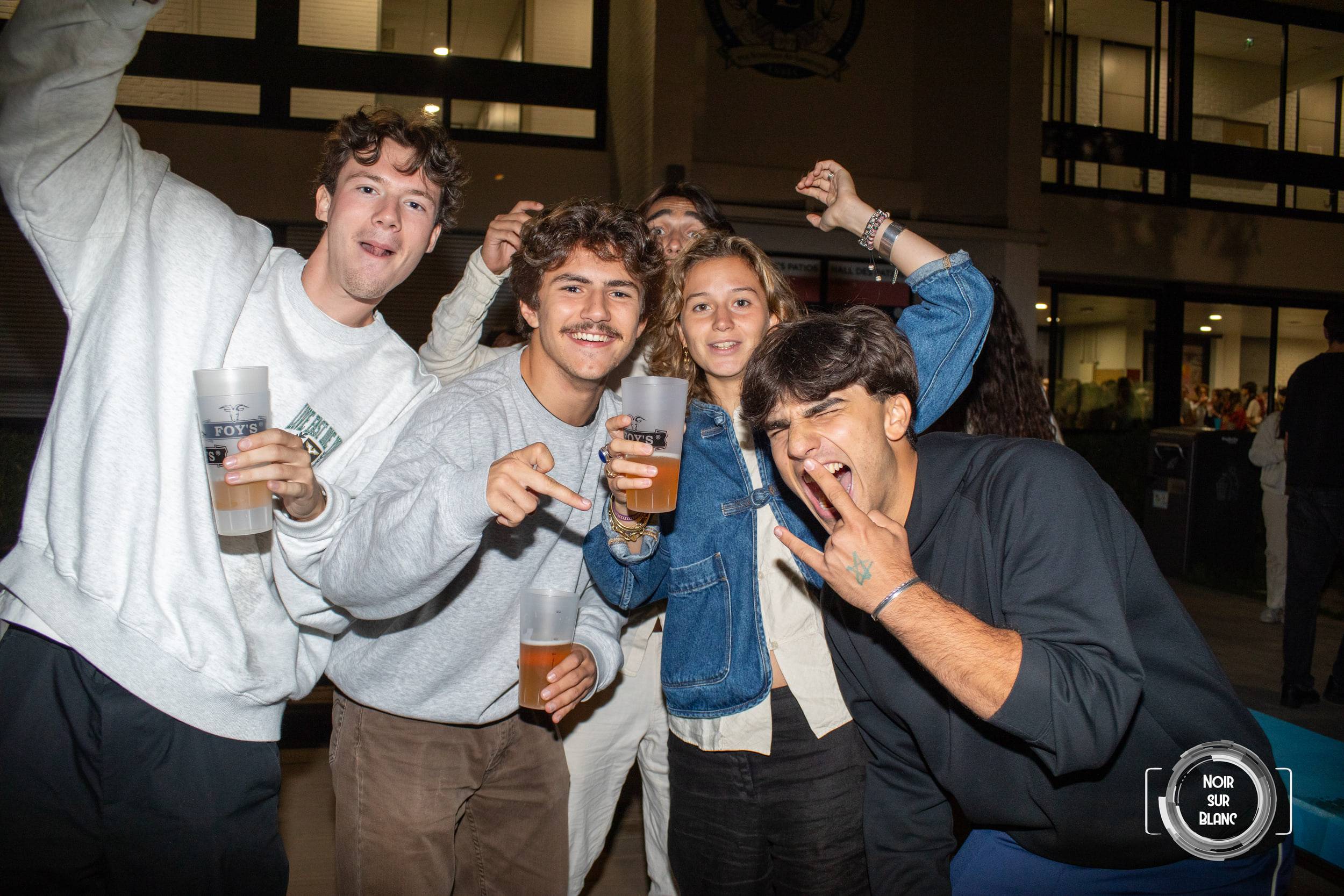 Cinq étudiants serrés les uns contre les autres brandissent des gobelets de bière vers l'objectif, sous un porche éclairé.