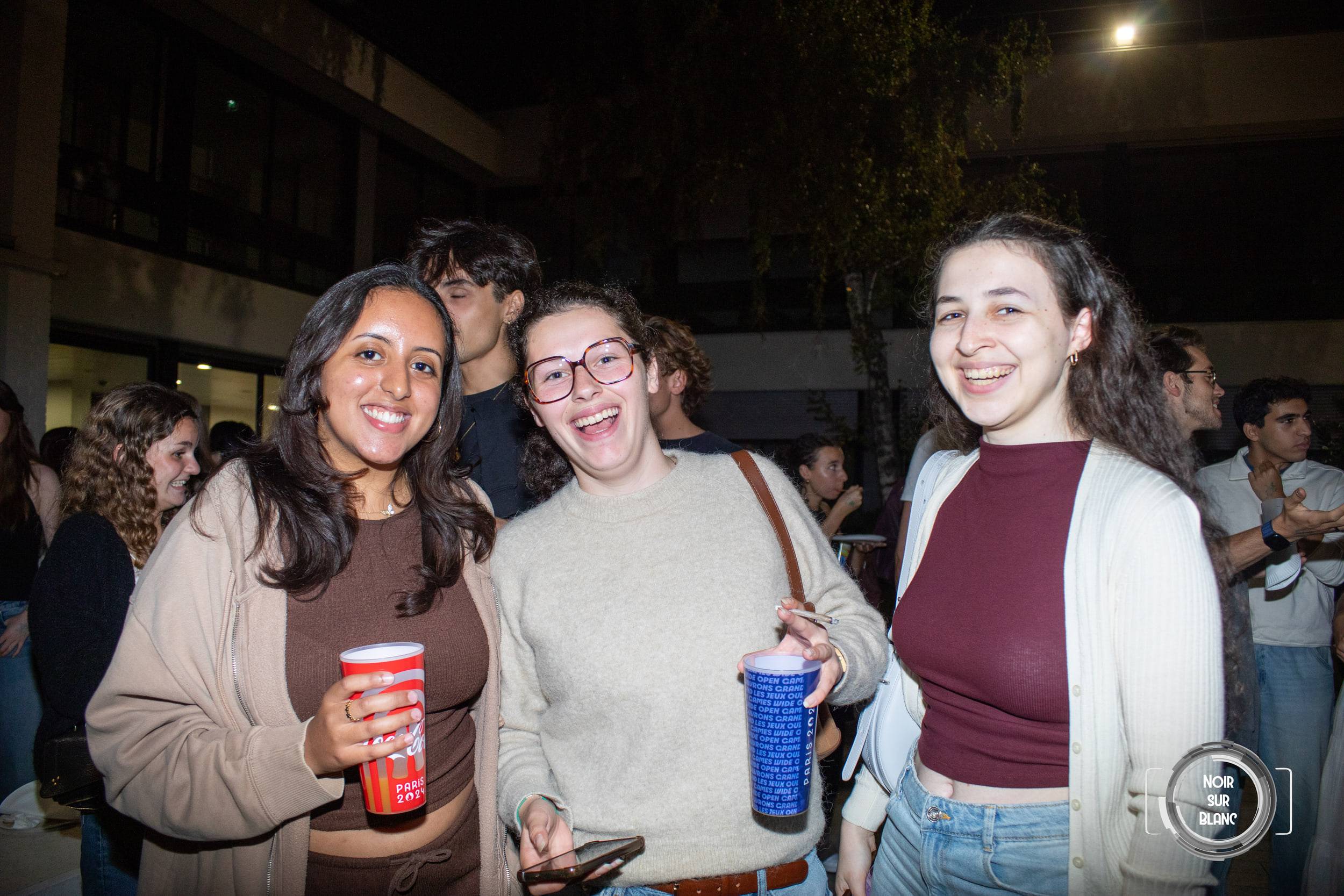 Trois étudiantes sourient face à l'objectif, gobelets rouge et bleu à la main, la foule et un bâtiment éclairé derrière elles.