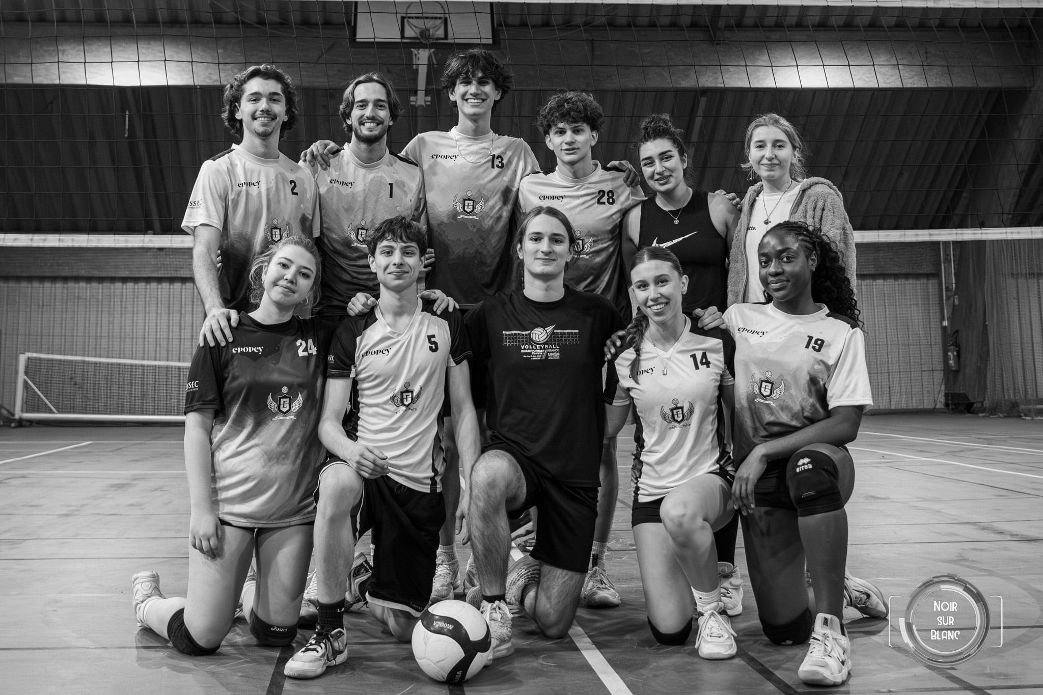 En noir et blanc, l'équipe de volley pose en deux rangées devant le filet, ballon posé au sol, à la fin du match.