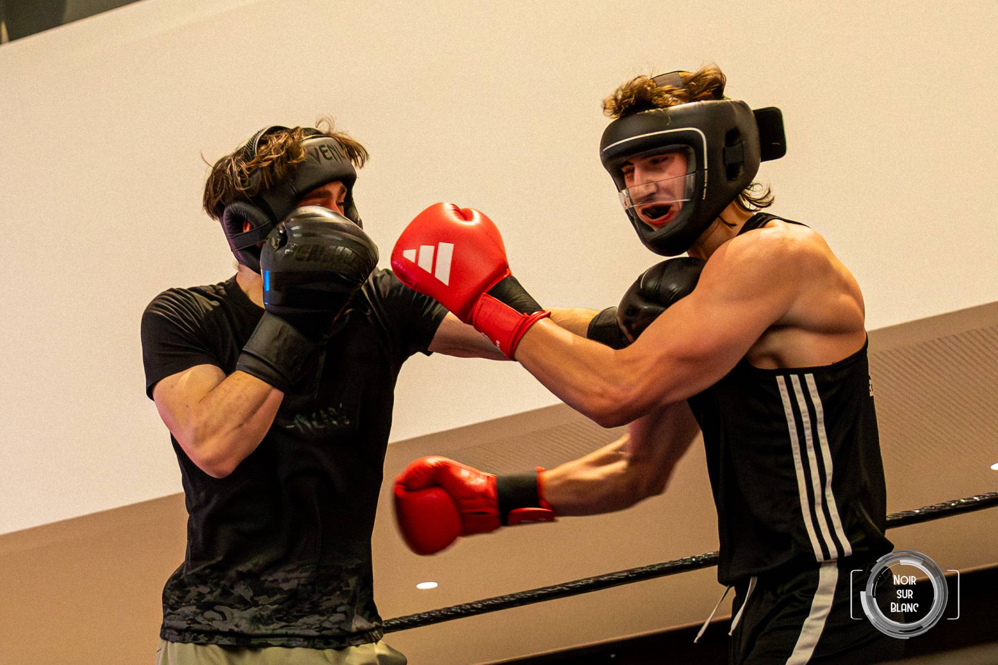 Deux boxeurs casqués échangent des coups sur le ring, l'un touchant l'autre au visage d'un gant rouge.
