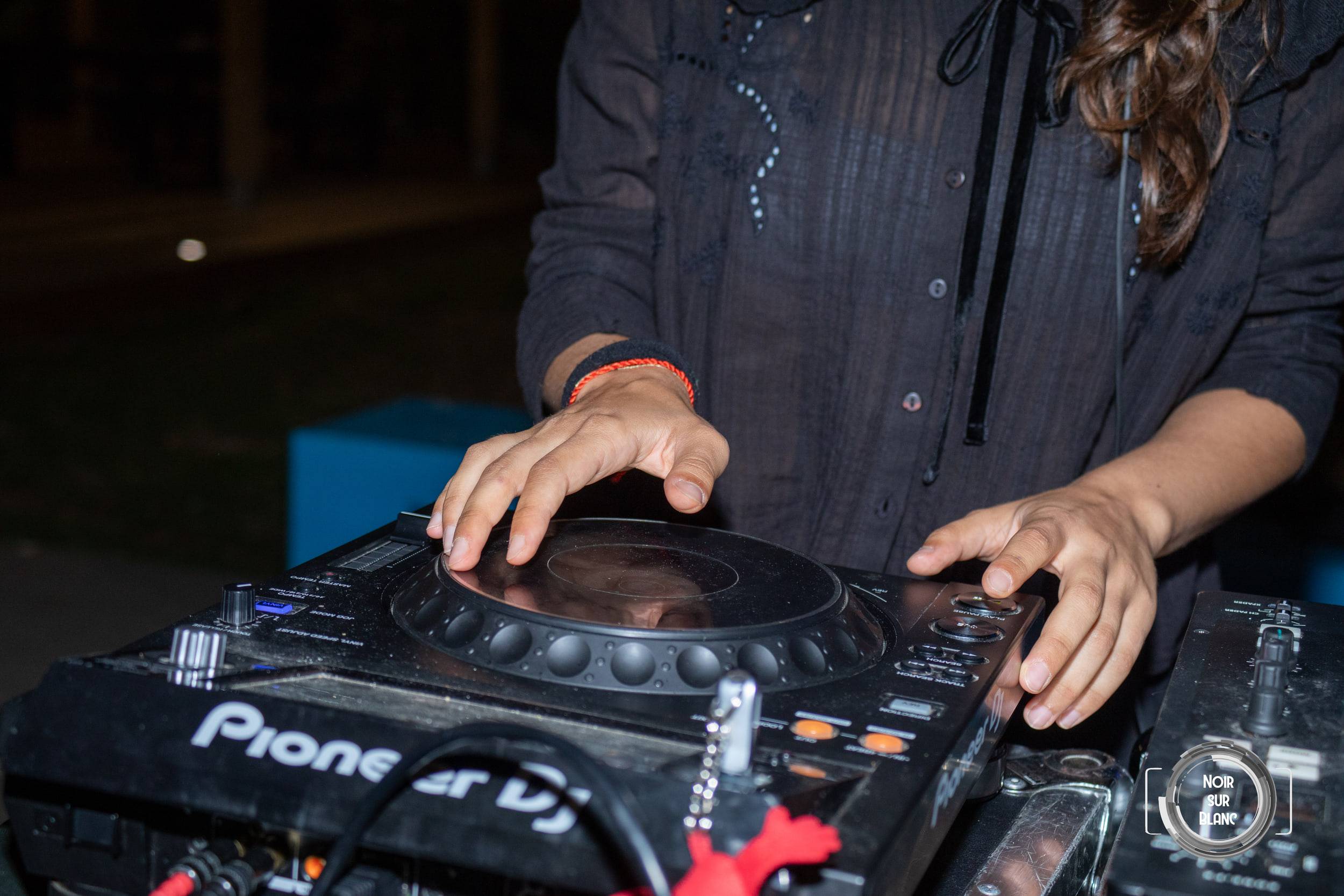 Gros plan sur les mains d'une DJ posées sur une platine Pioneer, le reste de la scène plongé dans le noir.