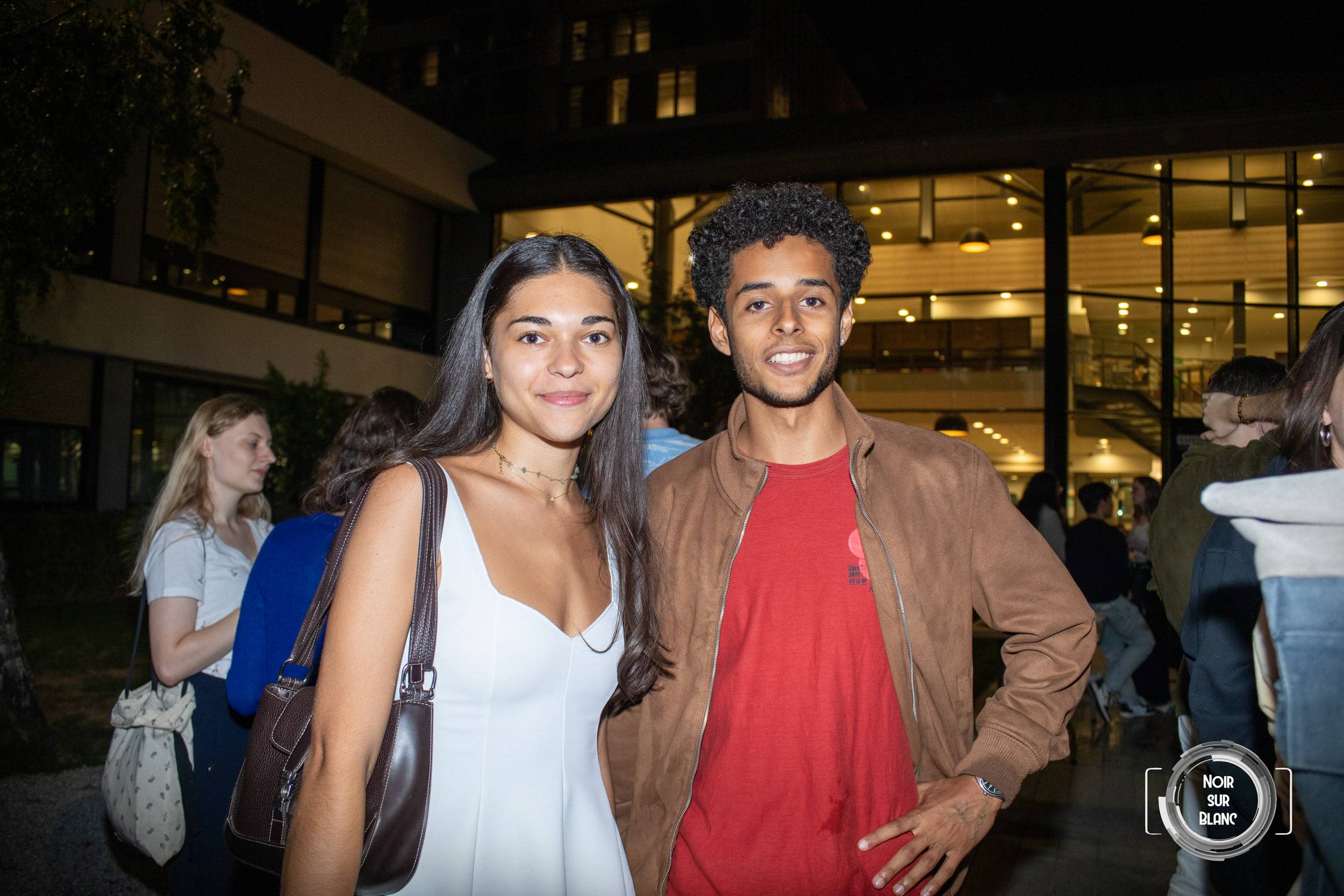 Une étudiante en robe blanche et un étudiant en t-shirt rouge posent devant le hall vitré illuminé, la soirée battant son plein derrière eux.