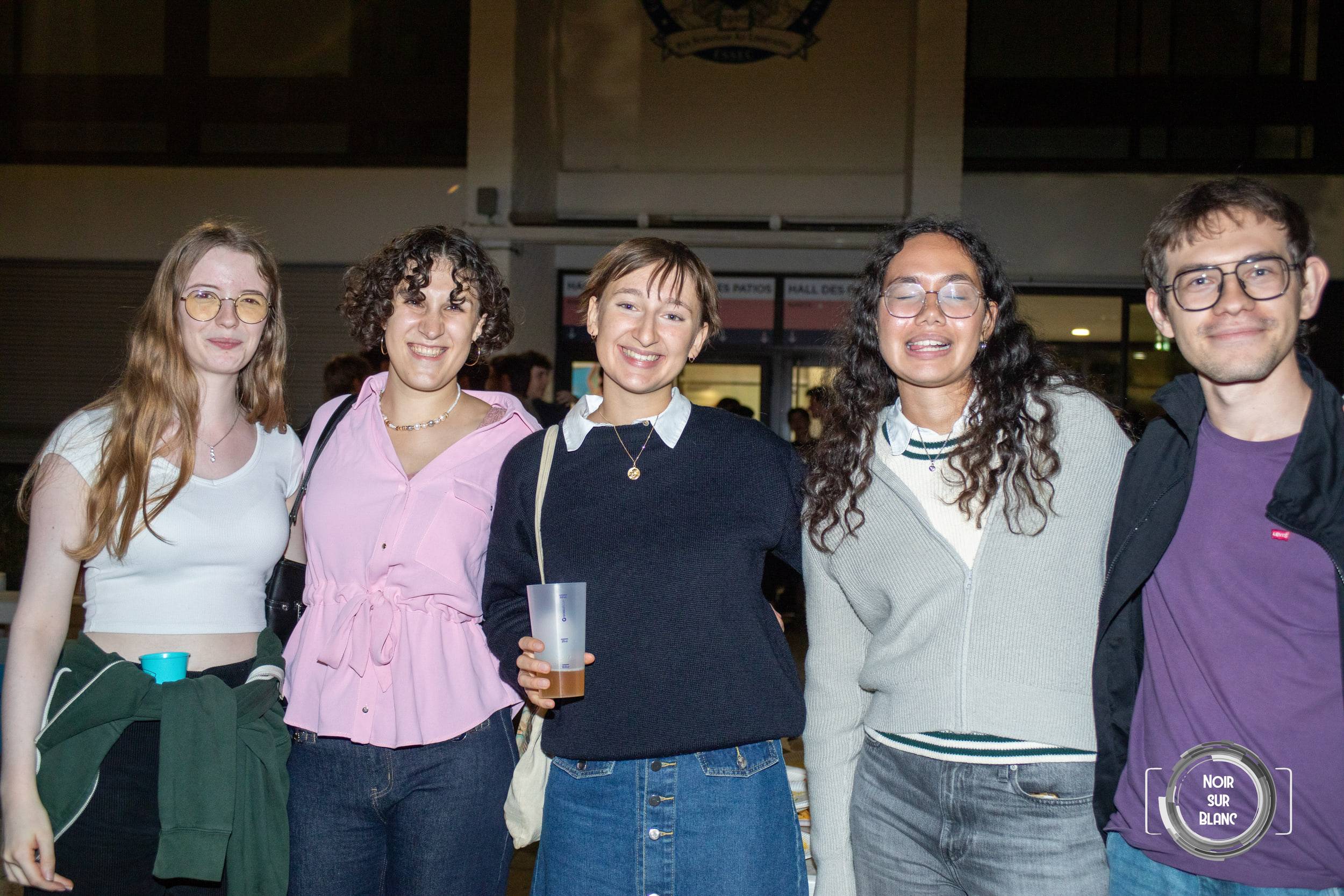 Cinq étudiants alignés sourient face à l'objectif devant l'entrée éclairée d'un bâtiment du campus.