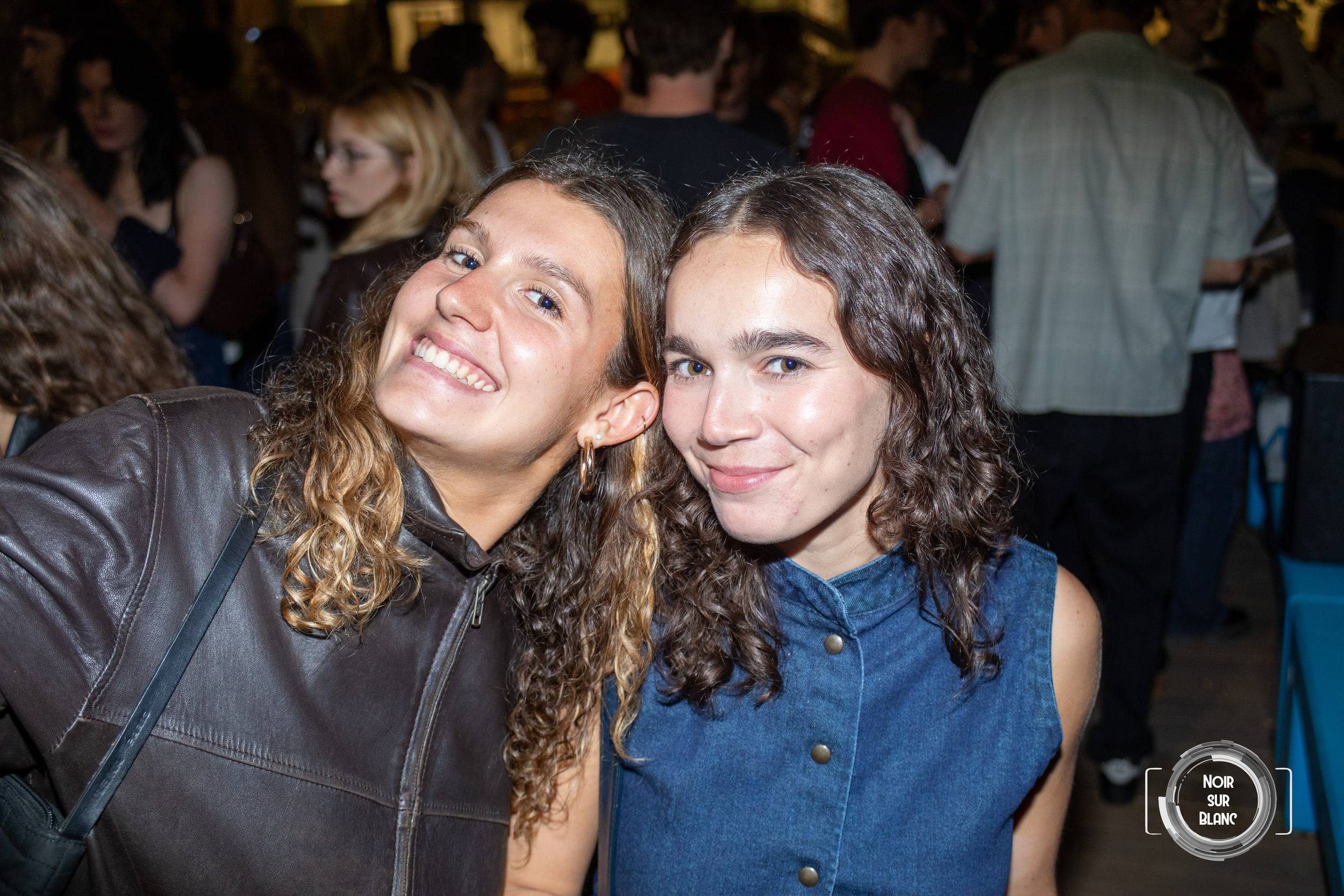 Deux amis penchent la tête l'un vers l'autre en souriant, la foule de la soirée floue derrière eux.