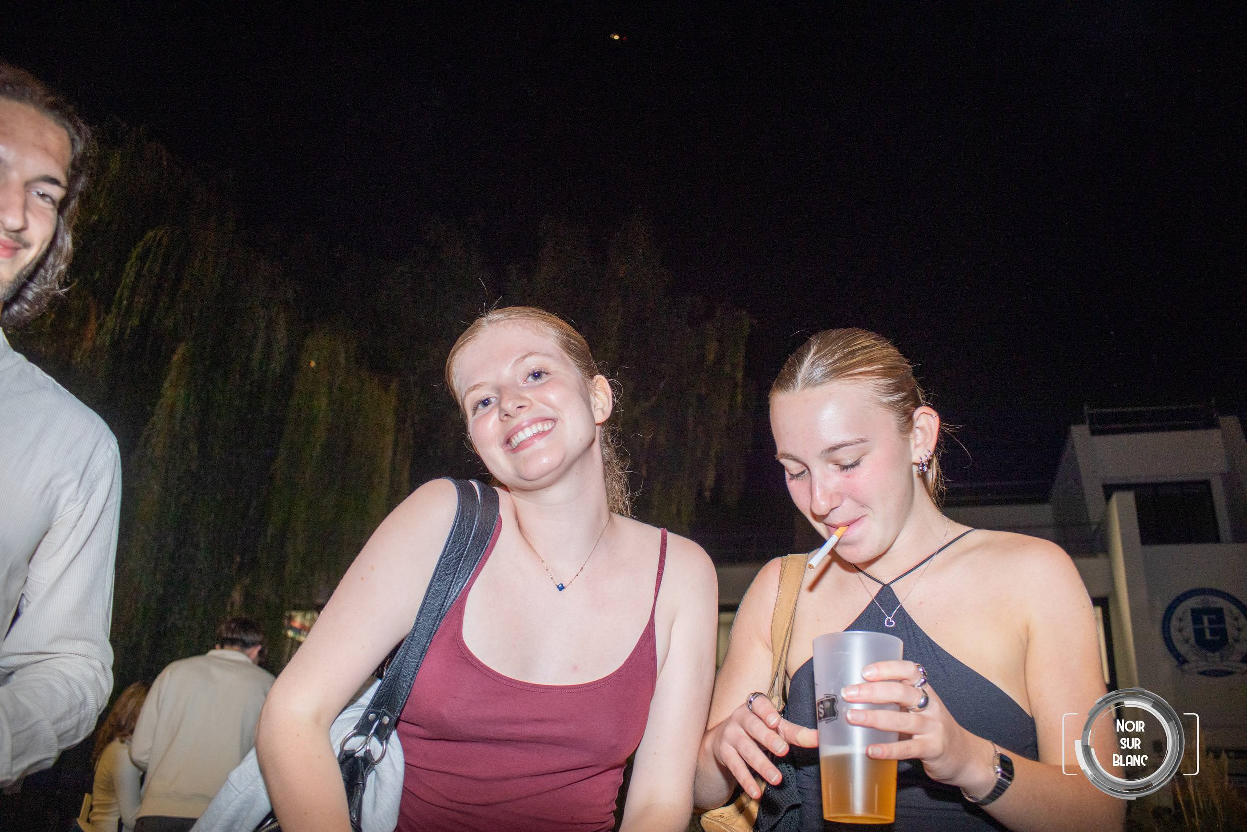 Deux étudiantes discutent dans la nuit, l'une boit à la paille dans un gobelet de bière, un saule éclairé derrière elles.