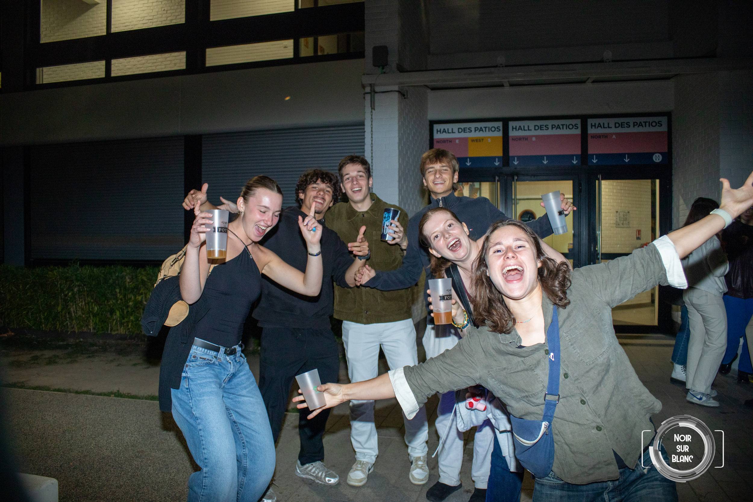 Un groupe d'étudiants pose bras écartés devant l'entrée du Hall des Patios, gobelets à la main.
