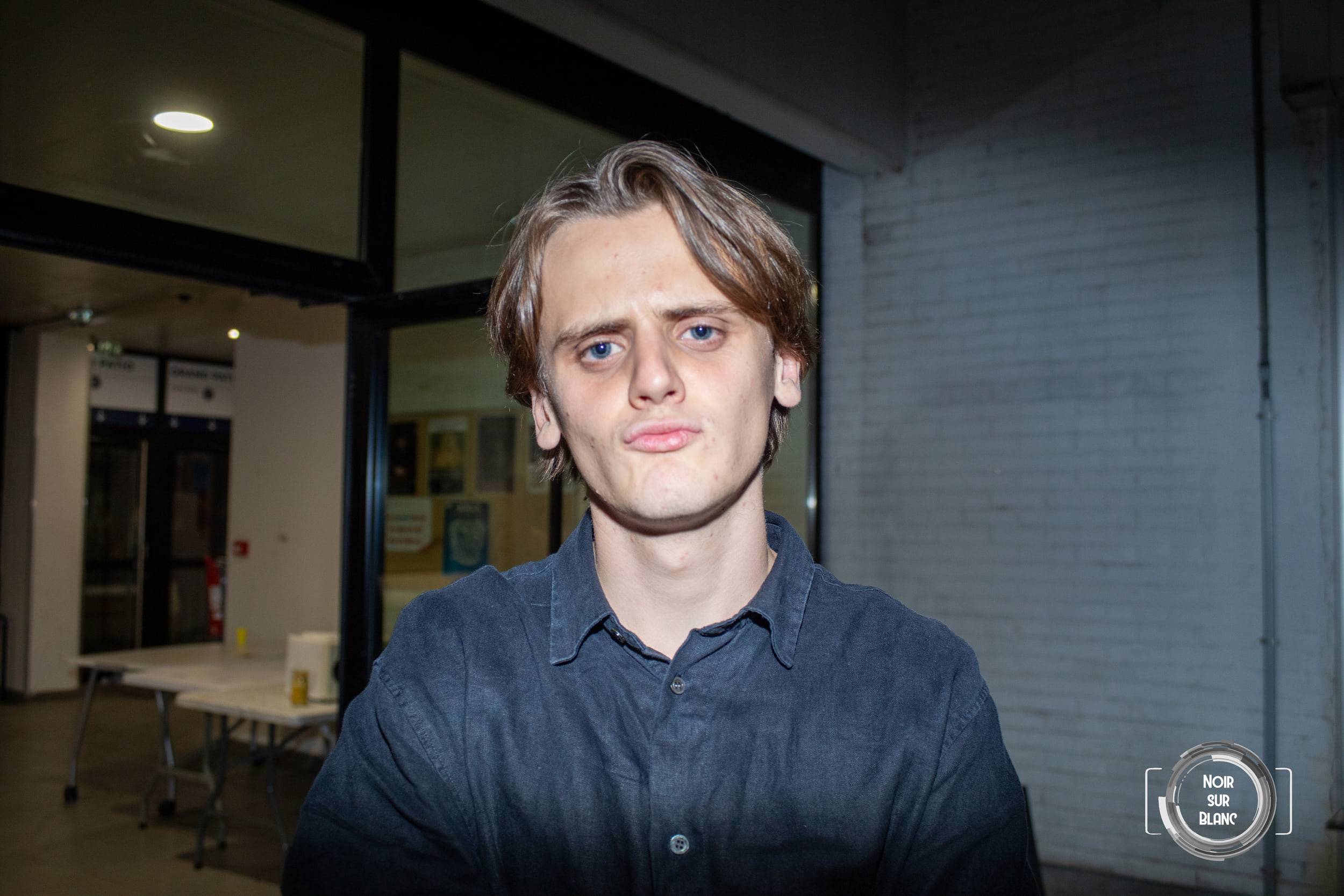 Portrait d'un étudiant en chemise sombre devant une salle vitrée éclairée, le regard droit vers l'objectif.