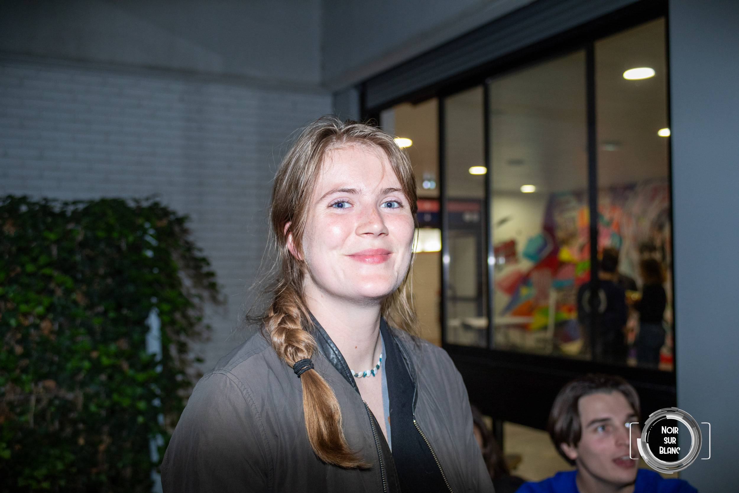 Portrait d'une étudiante à la tresse, souriante, devant une baie vitrée et un mur couvert de lierre.