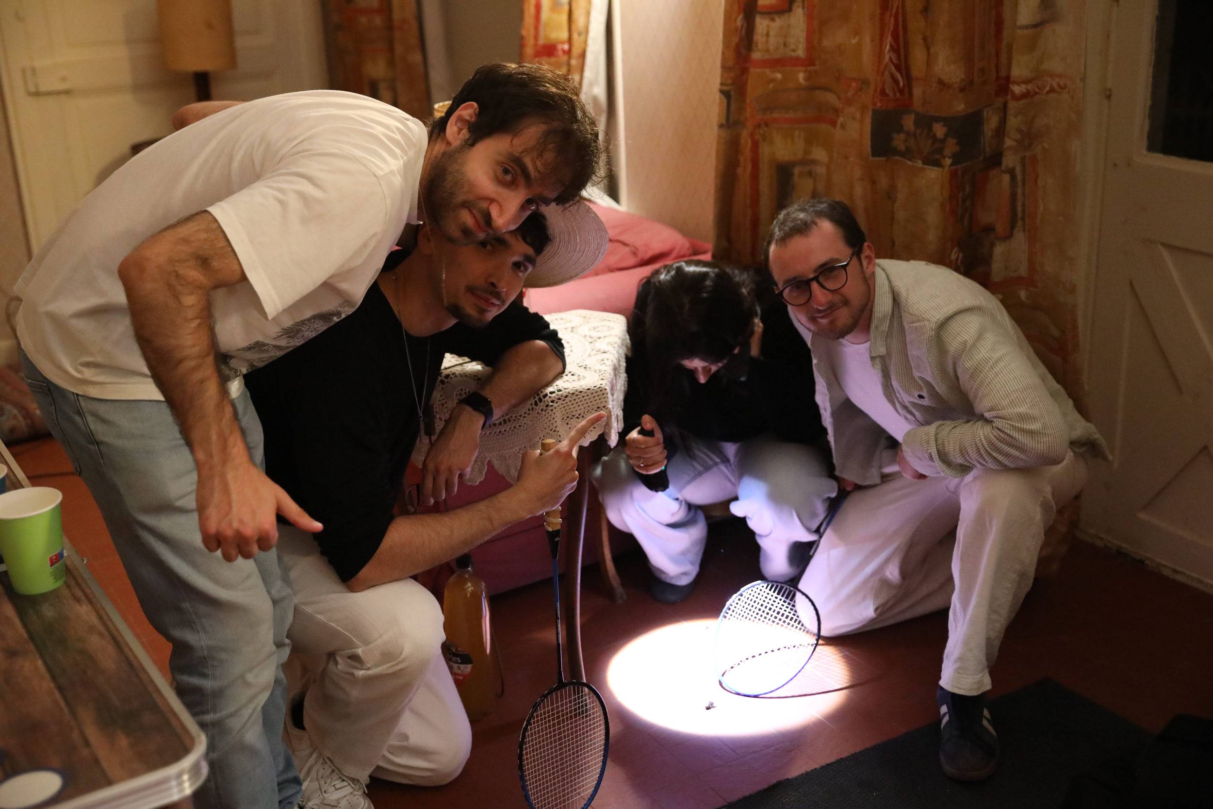 Quatre membres accroupis autour d'une lampe posée au sol et de raquettes de badminton, dans un salon aux boiseries peintes.
