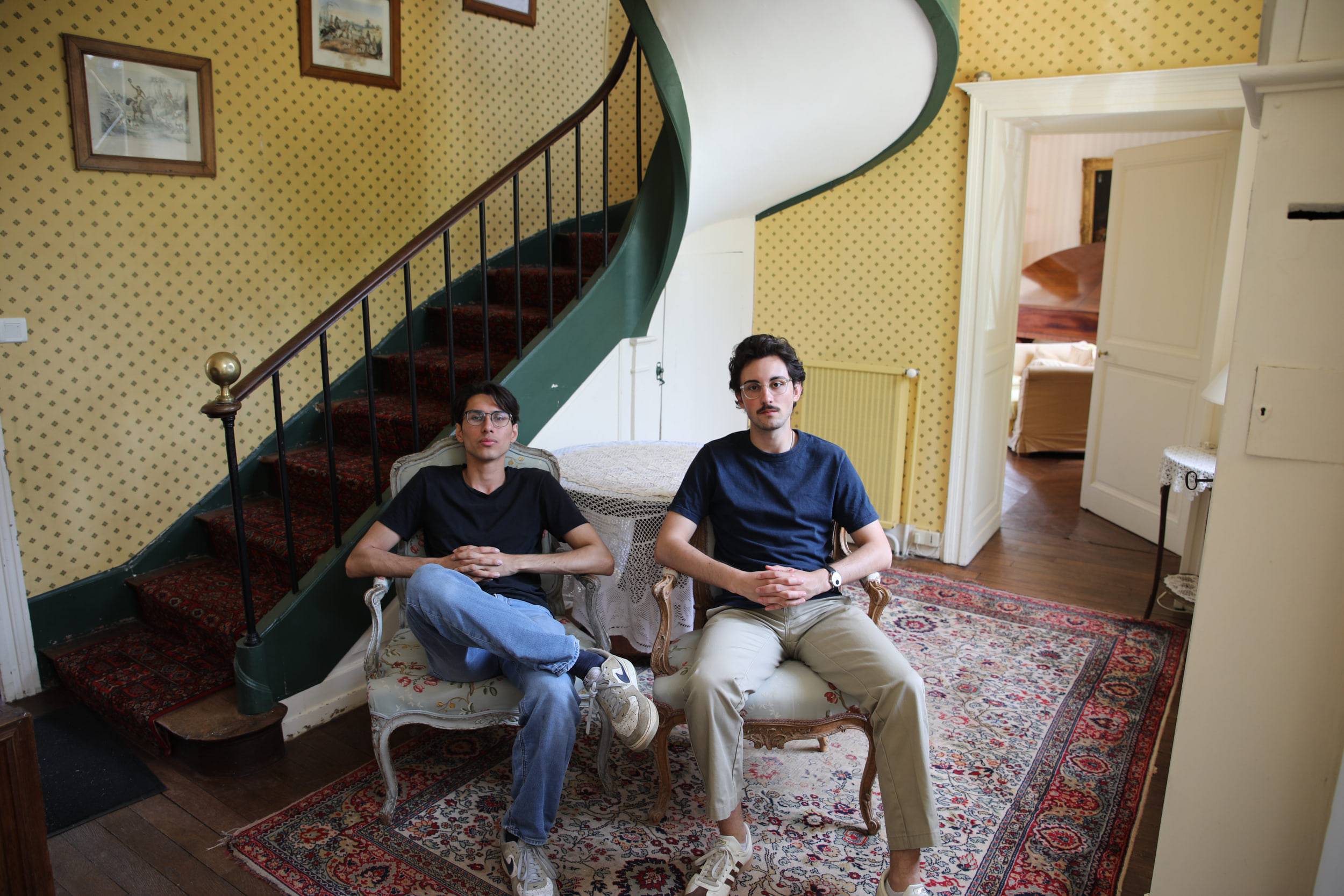 Deux membres assis dans un canapé et un fauteuil au pied d'un escalier tournant, dans un vestibule aux murs jaunes et au tapis persan.