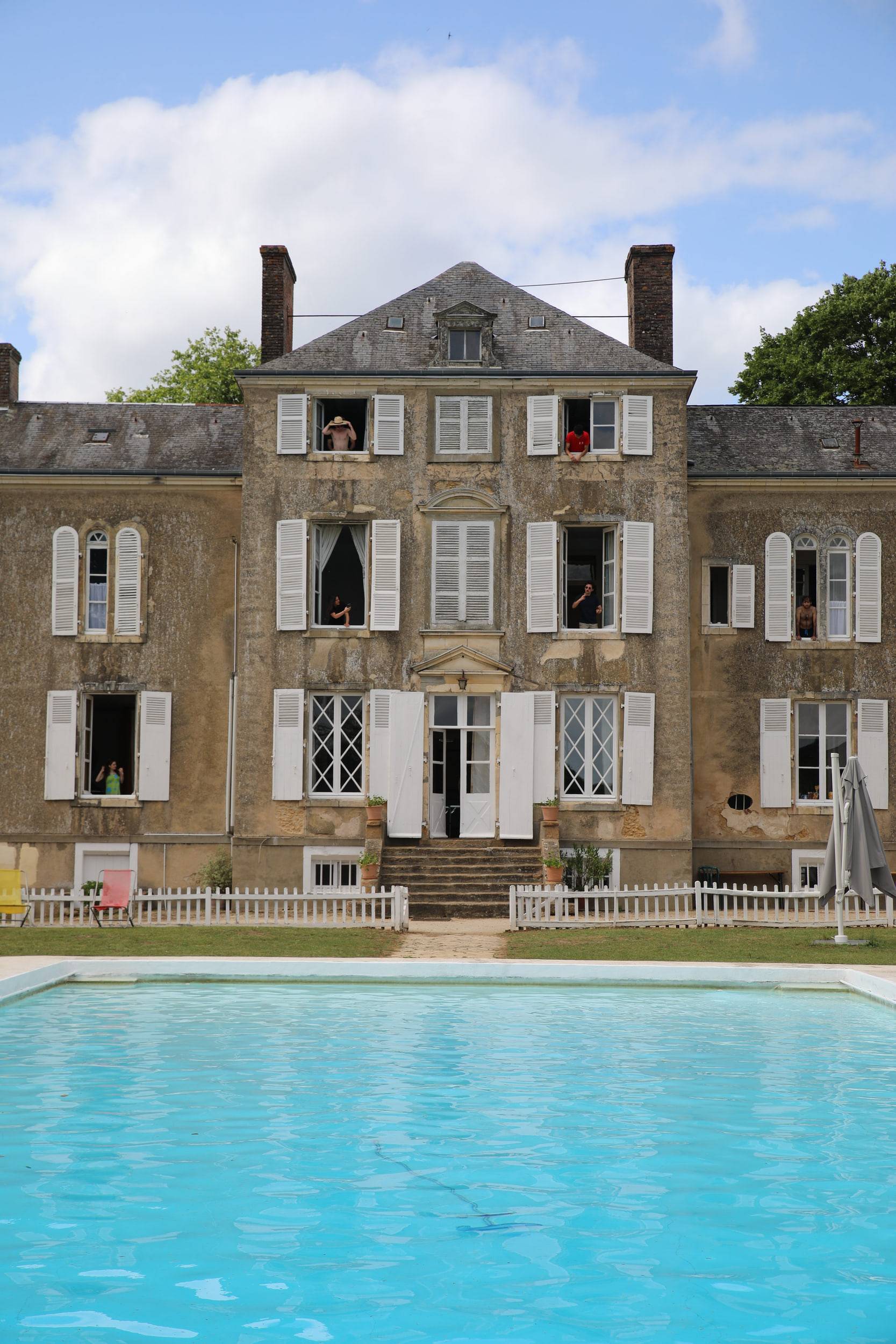 Façade d'une maison de maître en pierre aux volets blancs, quelques visages aux fenêtres, une piscine turquoise au premier plan.