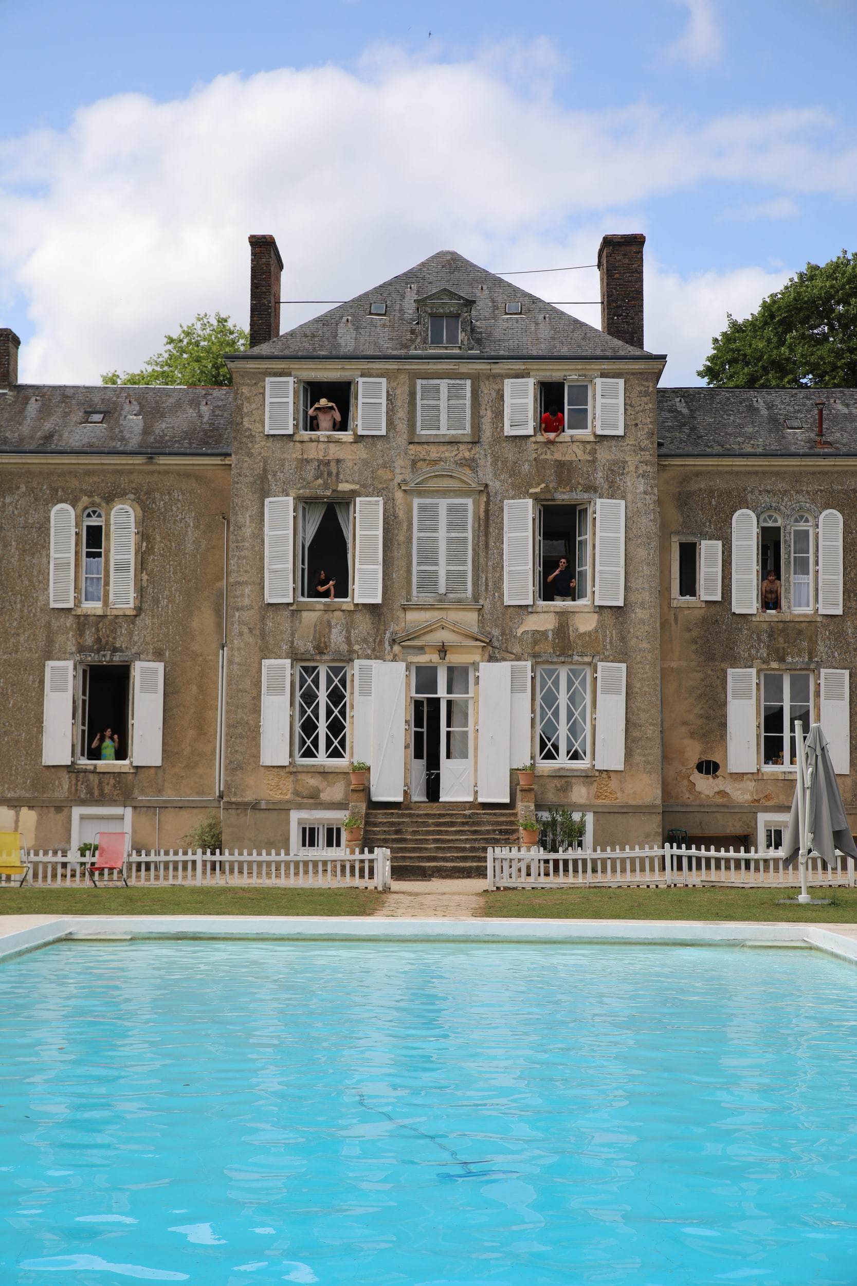 Façade d'une maison de maître en pierre aux volets blancs, quelques visages aux fenêtres, une piscine turquoise au premier plan.