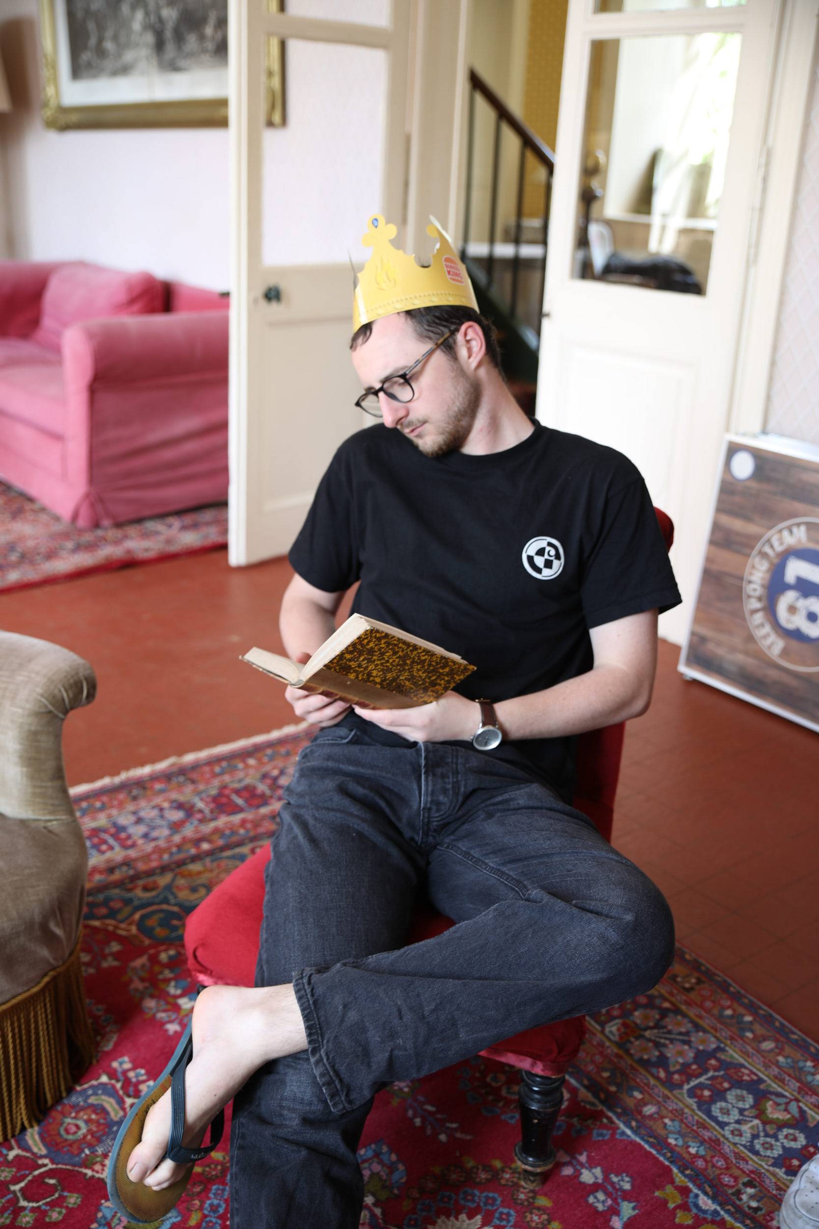 Un membre coiffé d'une couronne en papier doré lit un livre, jambes croisées dans un fauteuil rouge posé sur un tapis persan.