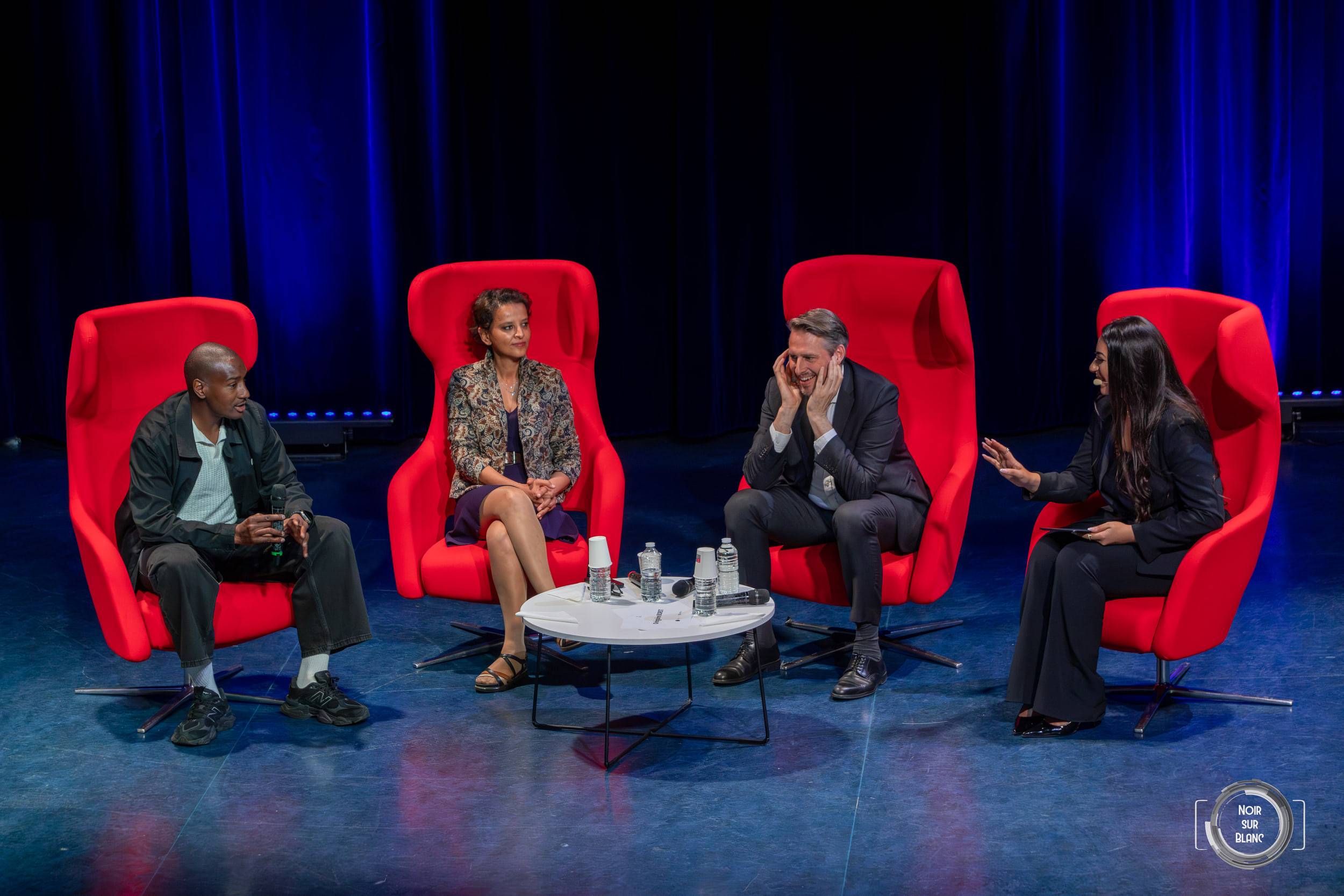 Quatre intervenants assis dans des fauteuils rouges autour d'une table basse, sur une scène tendue de bleu.