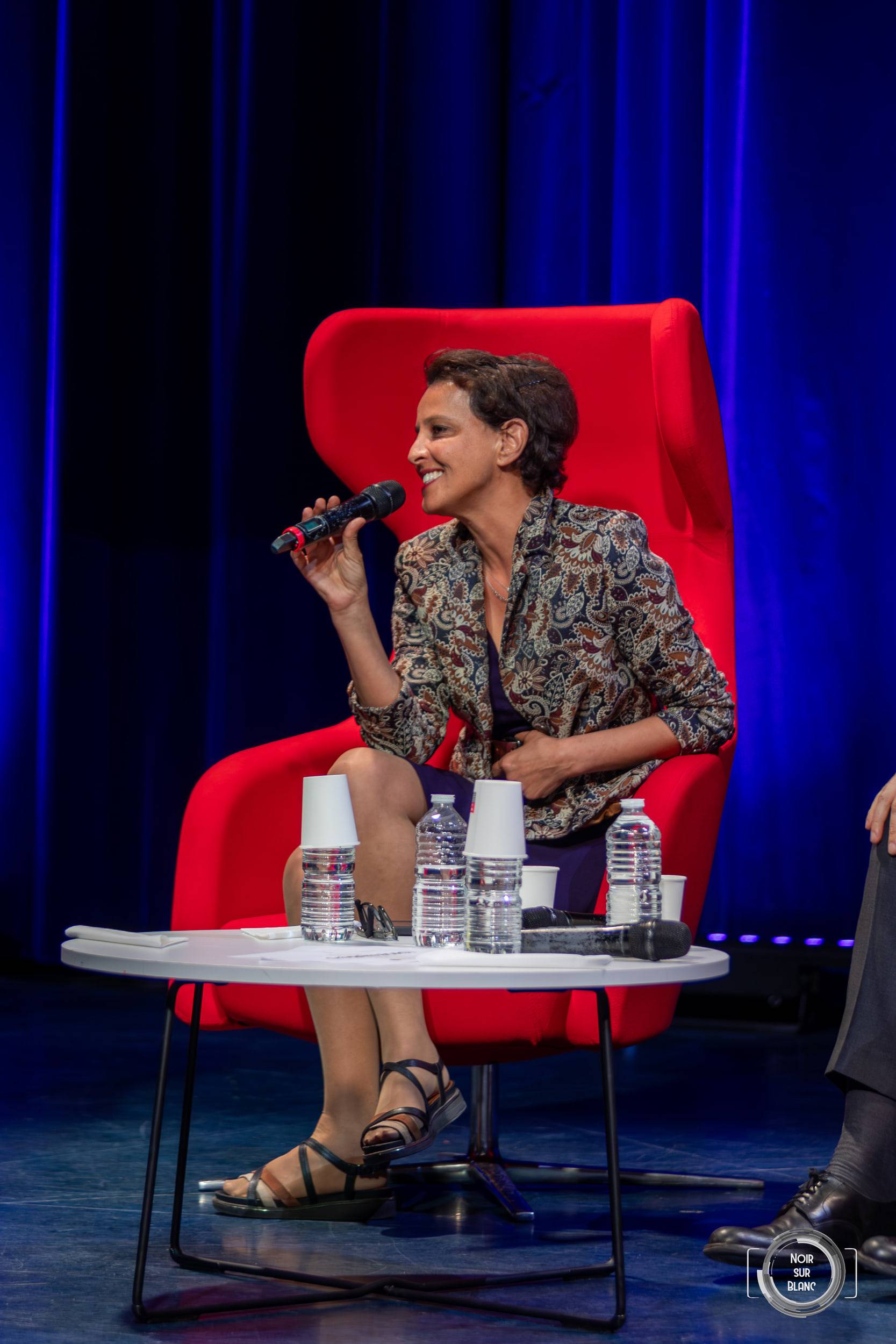 Une intervenante parle au micro depuis son fauteuil rouge, devant un rideau bleu éclairé.