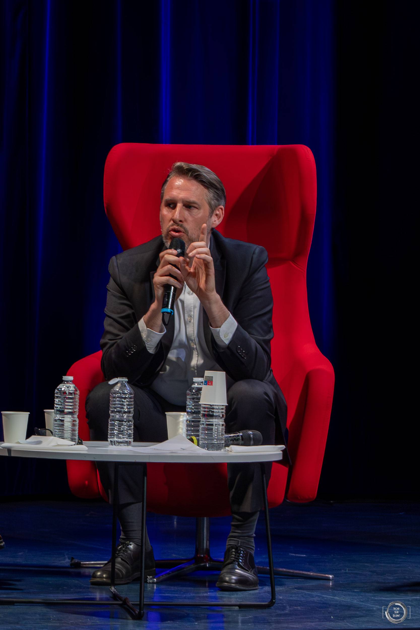 Un intervenant en costume sombre répond au micro, assis dans un fauteuil rouge sur la scène.