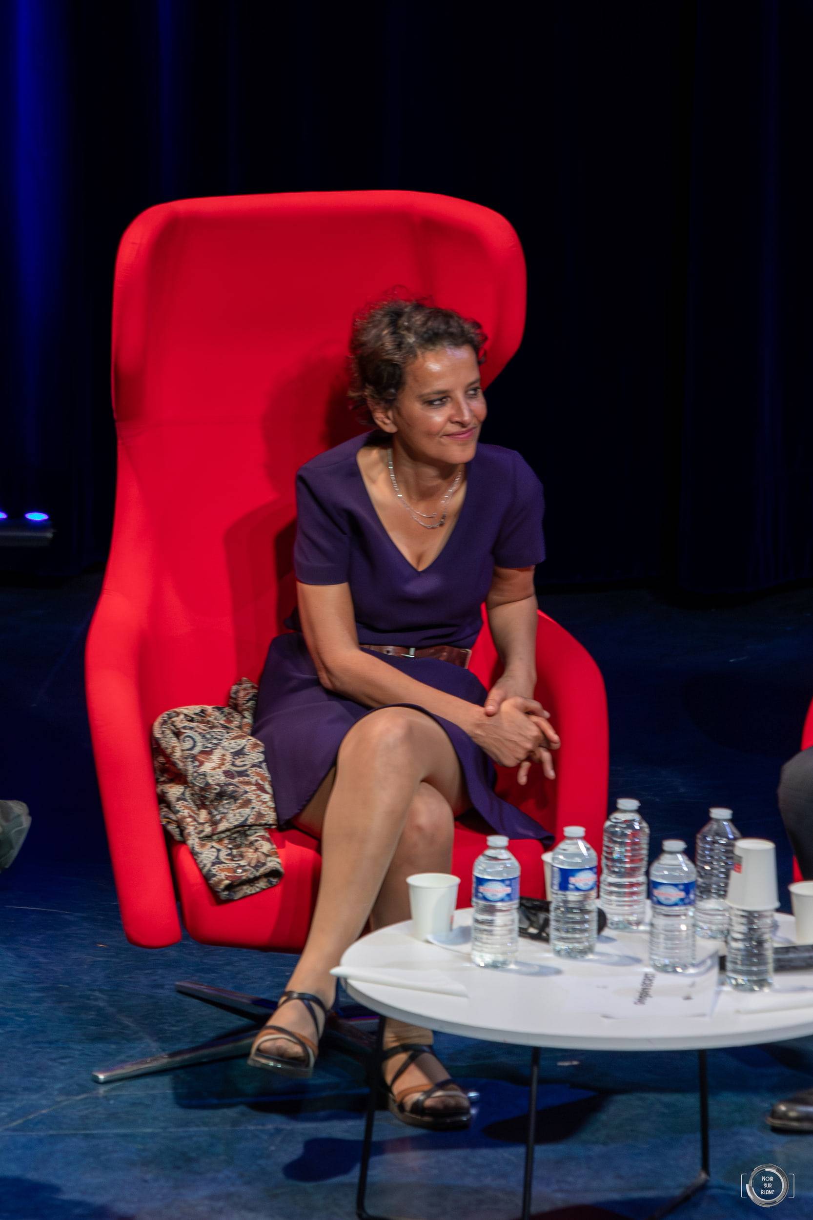 Une intervenante en robe violette, assise dans un fauteuil rouge, écoute les mains posées sur les genoux.
