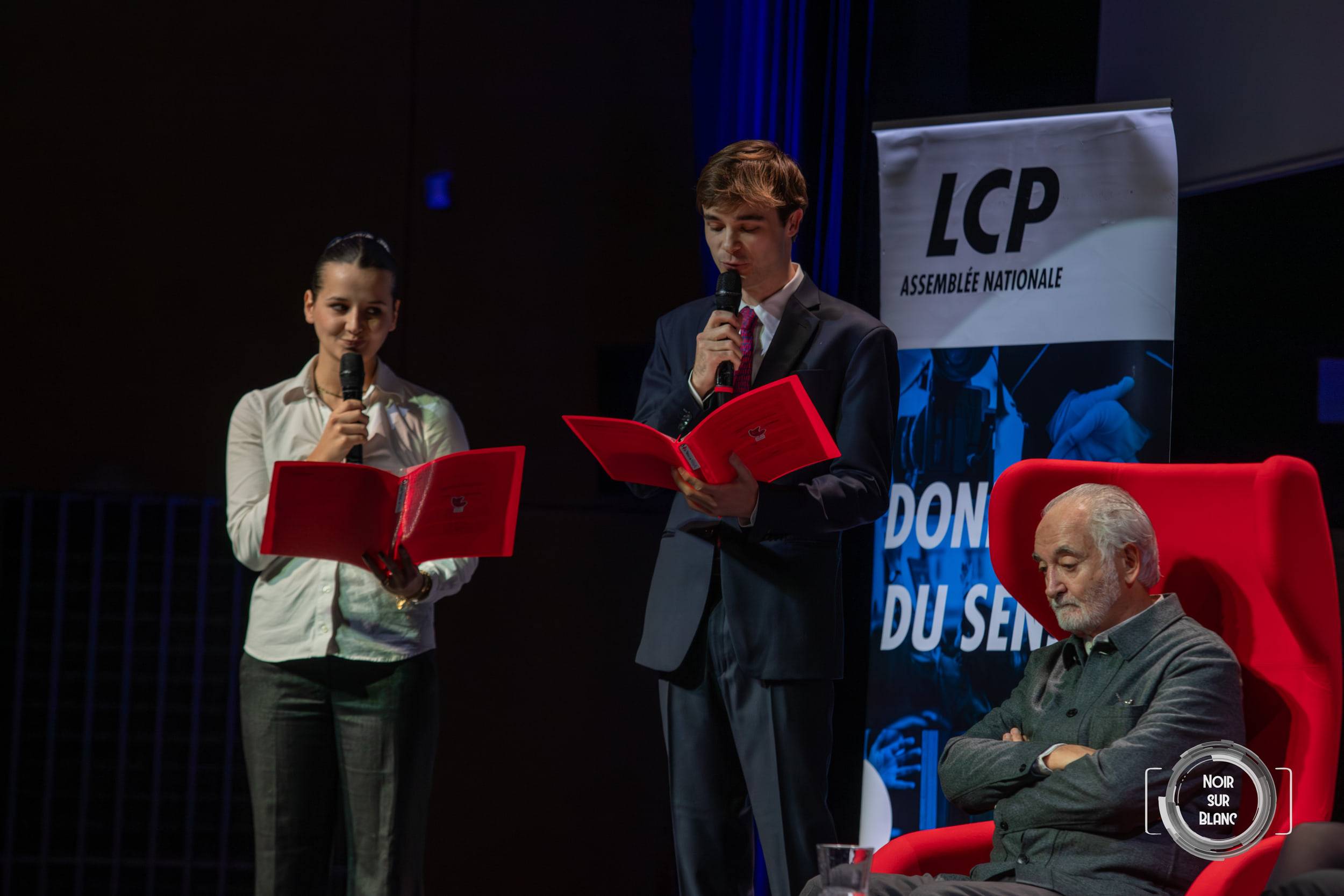 Deux étudiants lisent leurs questions dans des chemises rouges, debout au micro, tandis que l'invité les écoute bras croisés dans un fauteuil rouge.