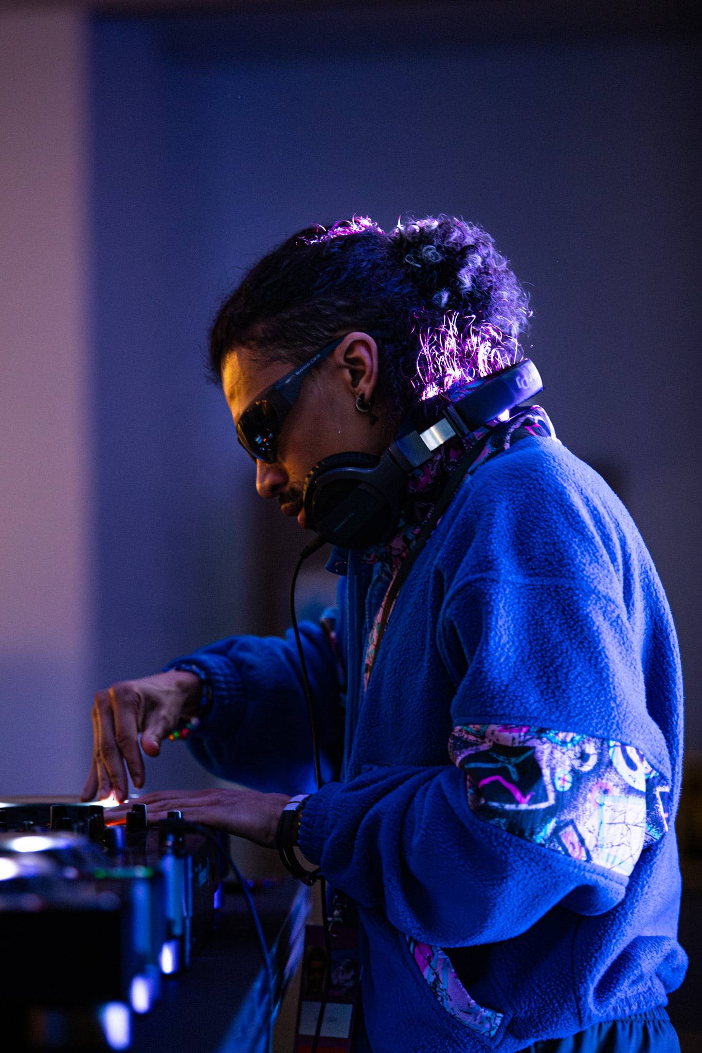 Un DJ en polaire bleue et lunettes de soleil, casque autour du cou, penché sur ses platines dans une lumière mauve.