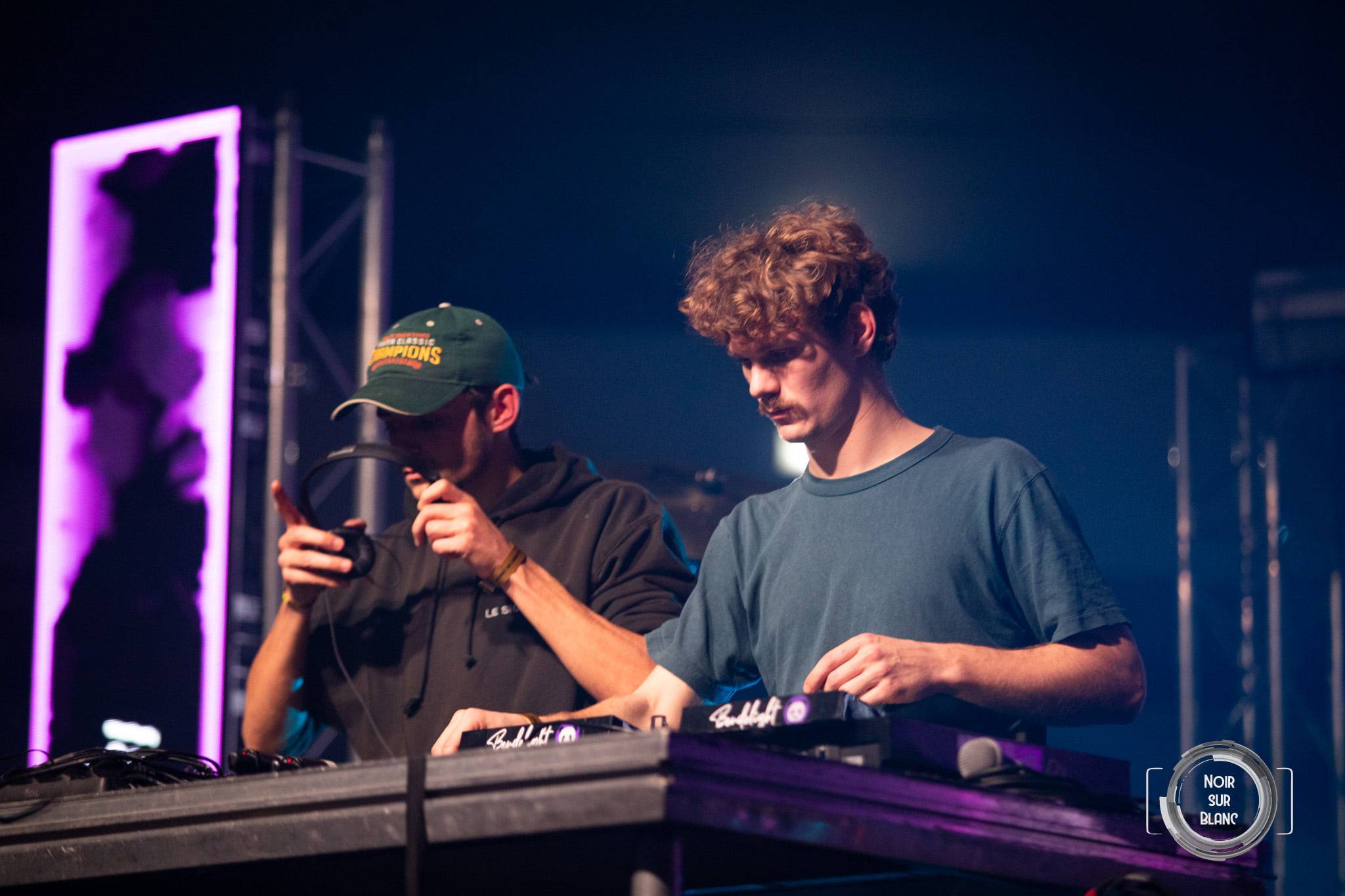 Deux DJ derrière les platines, l'un en casquette verte penché sur la table de mixage, devant un totem de lumière rose.
