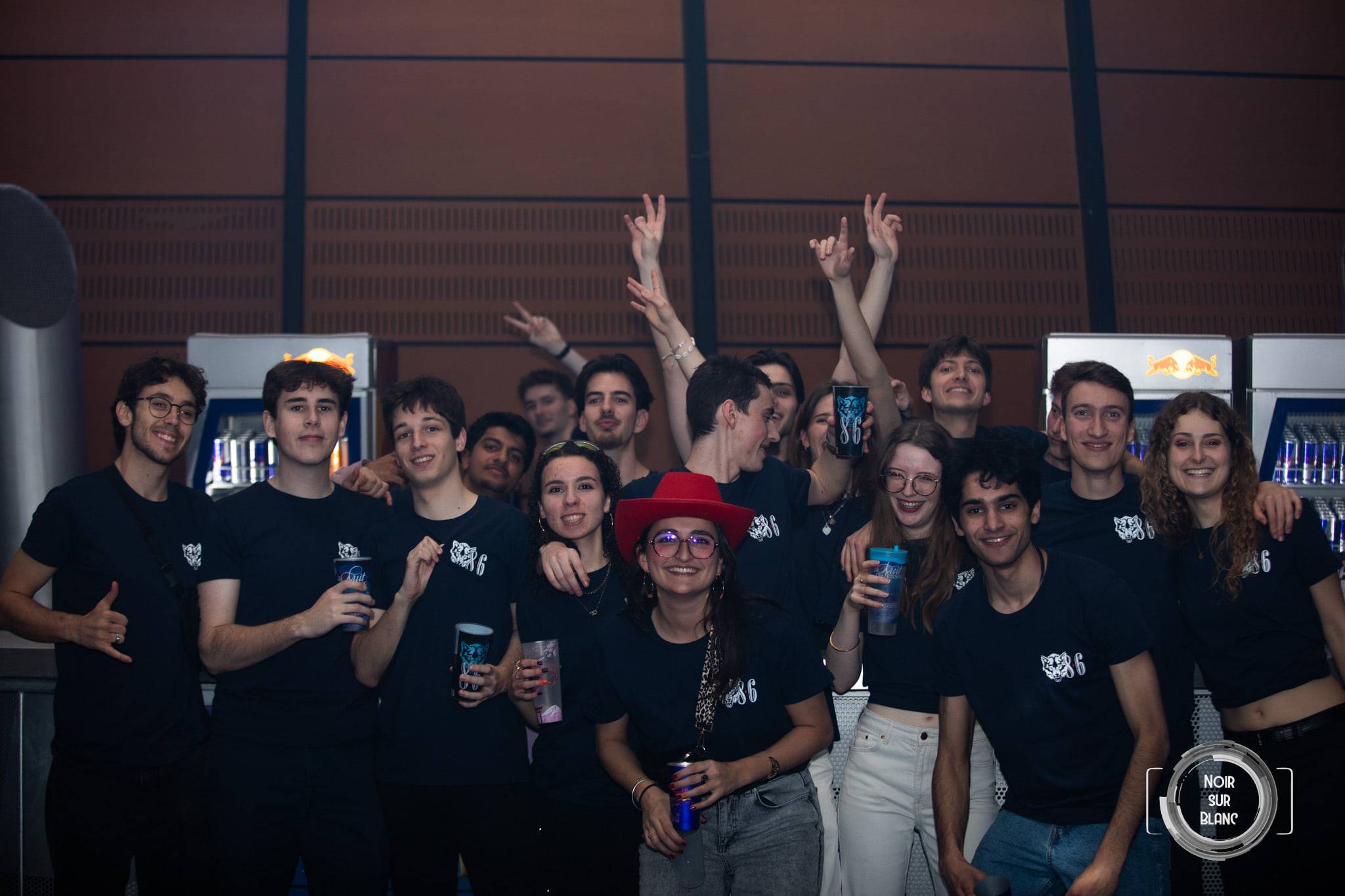 Une quinzaine d'étudiants en t-shirt noir posent bras levés devant les frigos d'un bar, gobelets à la main.