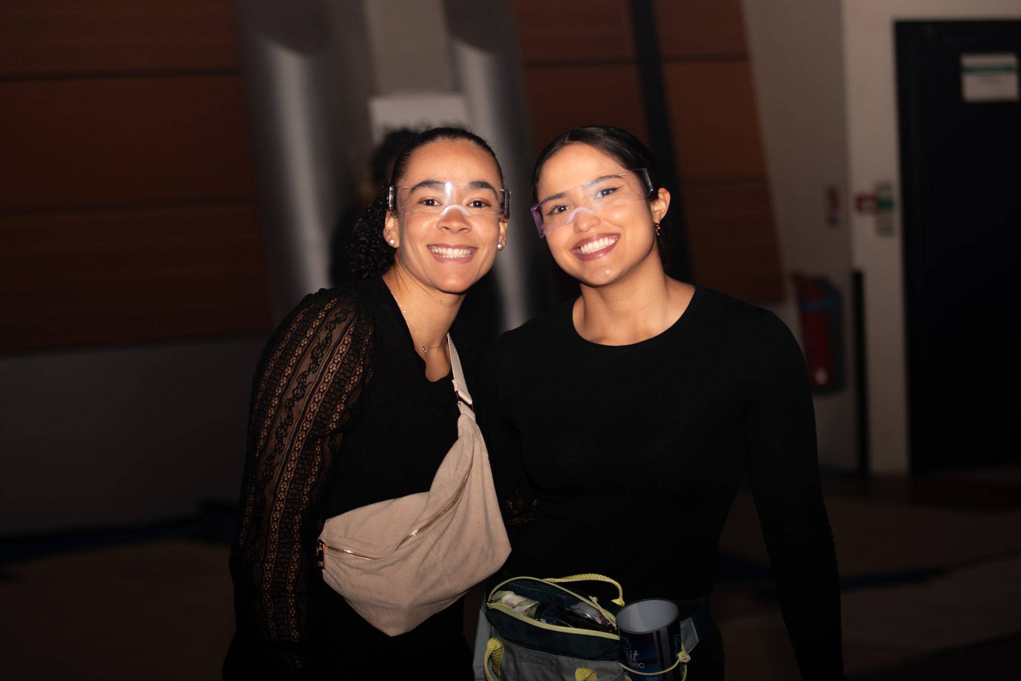 Deux étudiantes en noir posent bras dessus bras dessous dans un couloir sombre, sacoche en bandoulière.