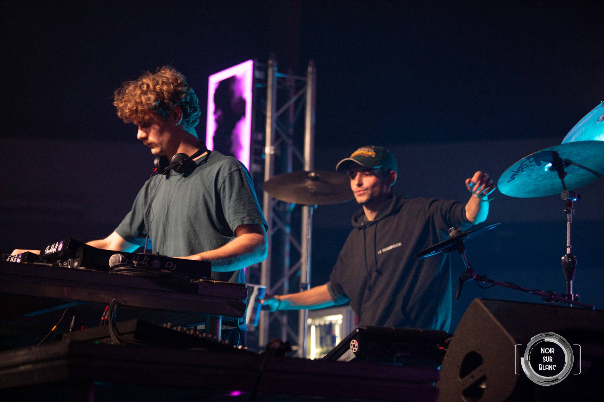 Un DJ mixe à gauche du cadre pendant qu'un batteur joue derrière lui, cymbales éclairées par un projecteur bleu.