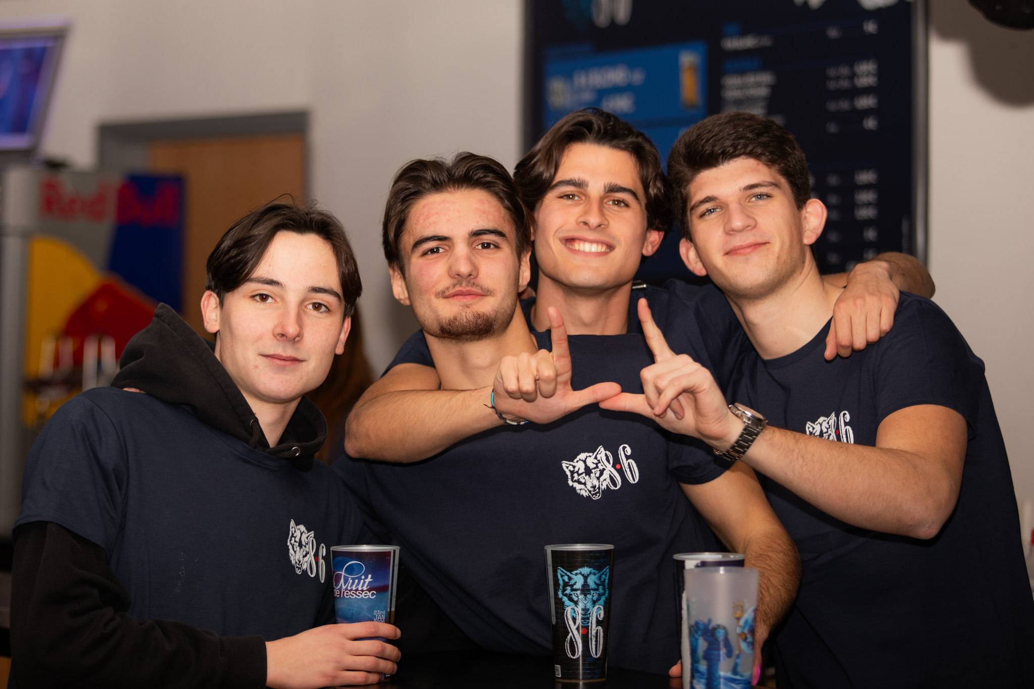 Quatre étudiants en sweat de promo posent bras autour des épaules, gobelets à la main, devant un stand Red Bull.