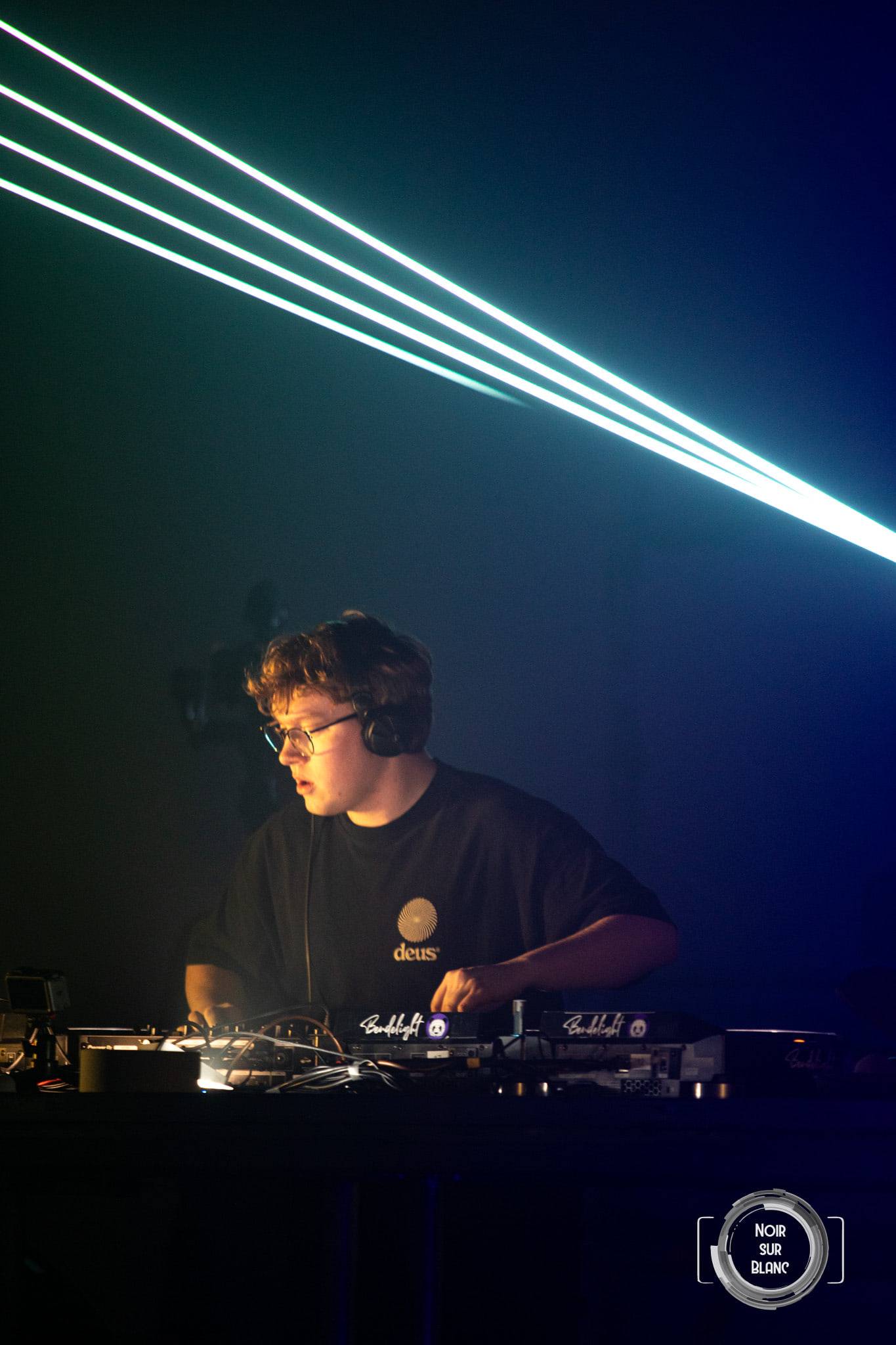 Quatre traits de laser turquoise traversent le haut du cadre au-dessus d'un DJ concentré sur ses platines.