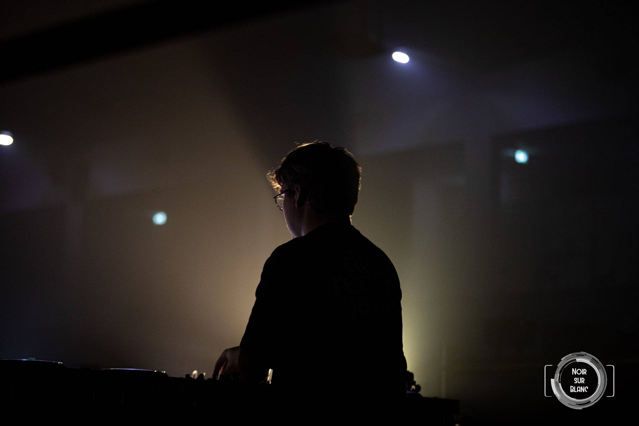 Un DJ de dos, en silhouette, devant un mur de fumée éclairé par un projecteur chaud.