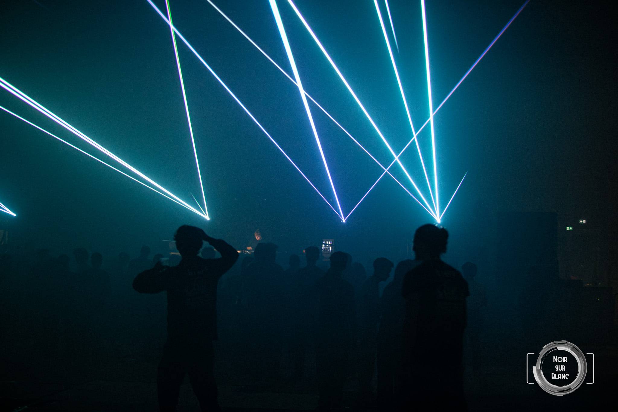 Des faisceaux laser turquoise se croisent au-dessus d'une piste où les danseurs ne sont plus que des silhouettes.