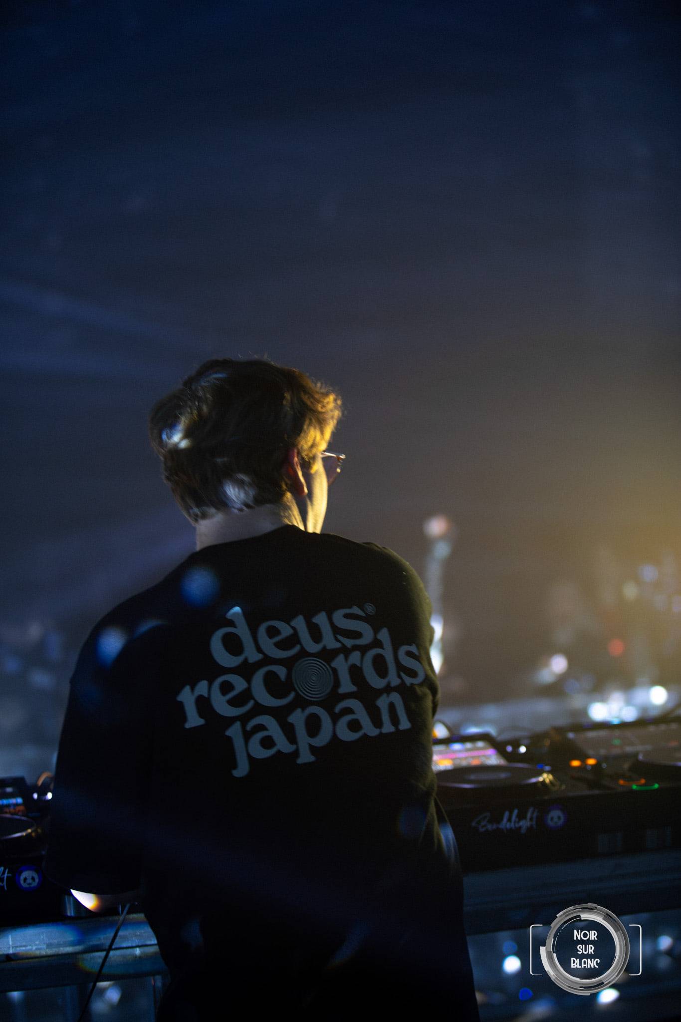 Un DJ de dos derrière ses platines, le dos de son blouson barré de l'inscription « deus records japan ».