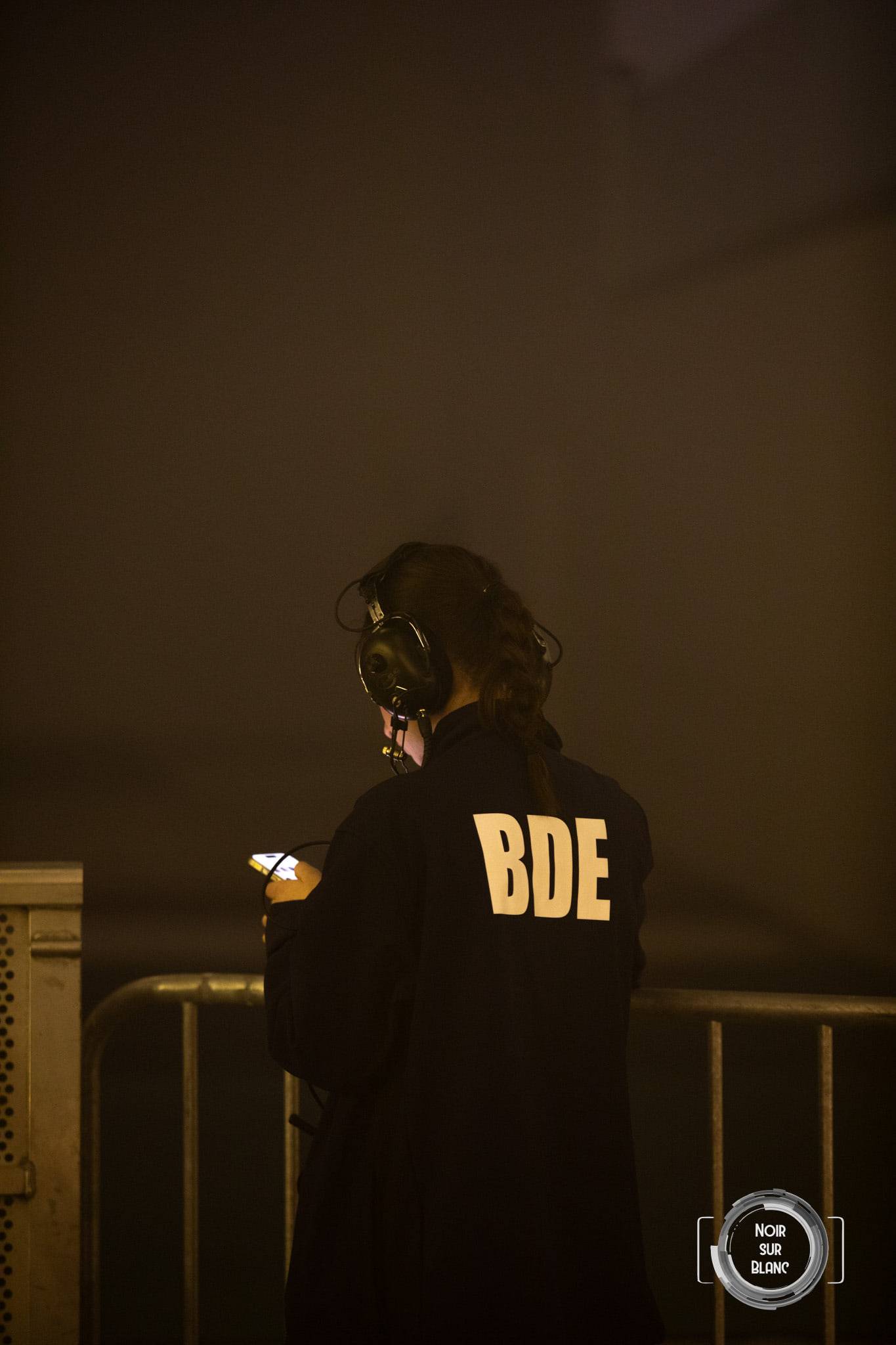 Une silhouette de dos accoudée à une barrière, casque autour du cou, les trois lettres BDE inscrites en doré dans le dos.