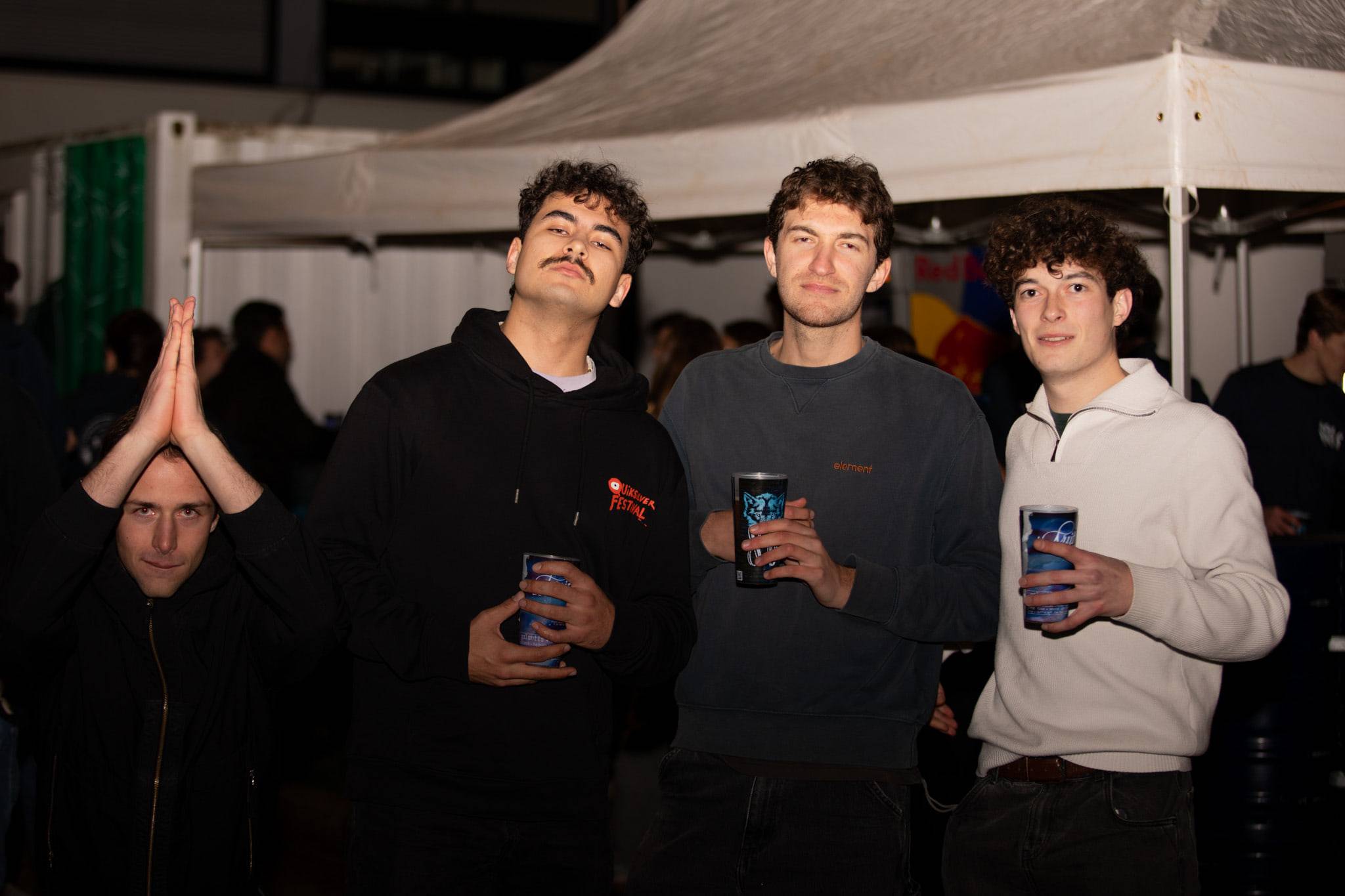 Quatre étudiants posent avec leurs gobelets devant les tentes blanches du village, l'un d'eux les mains jointes au-dessus de la tête.