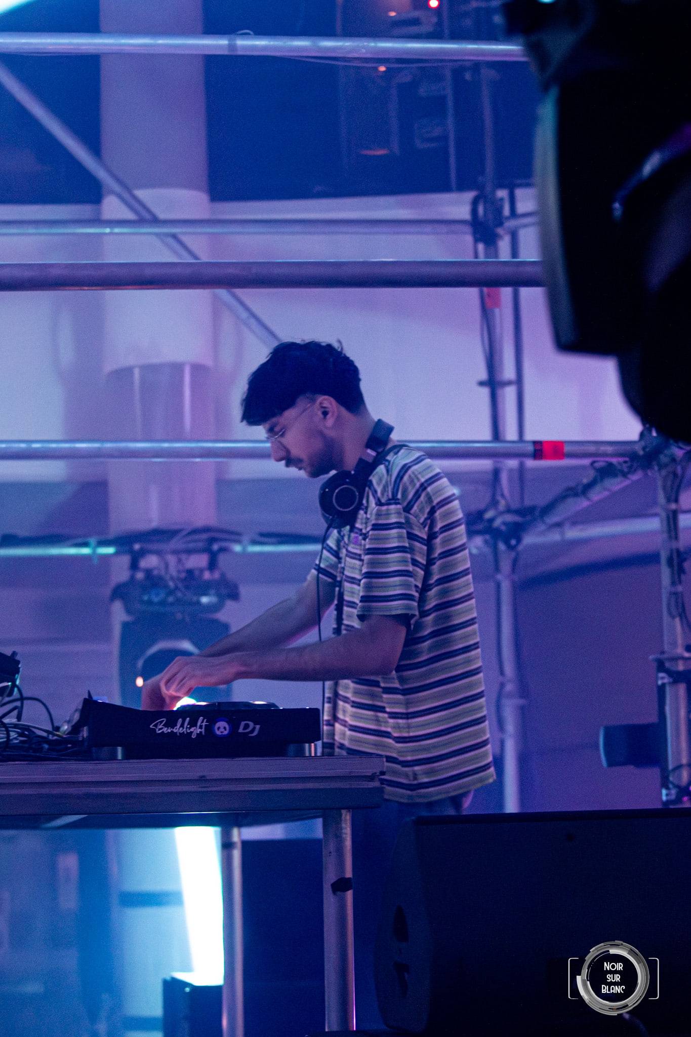 Un DJ en polo rayé mixe sous une structure métallique baignée de violet.