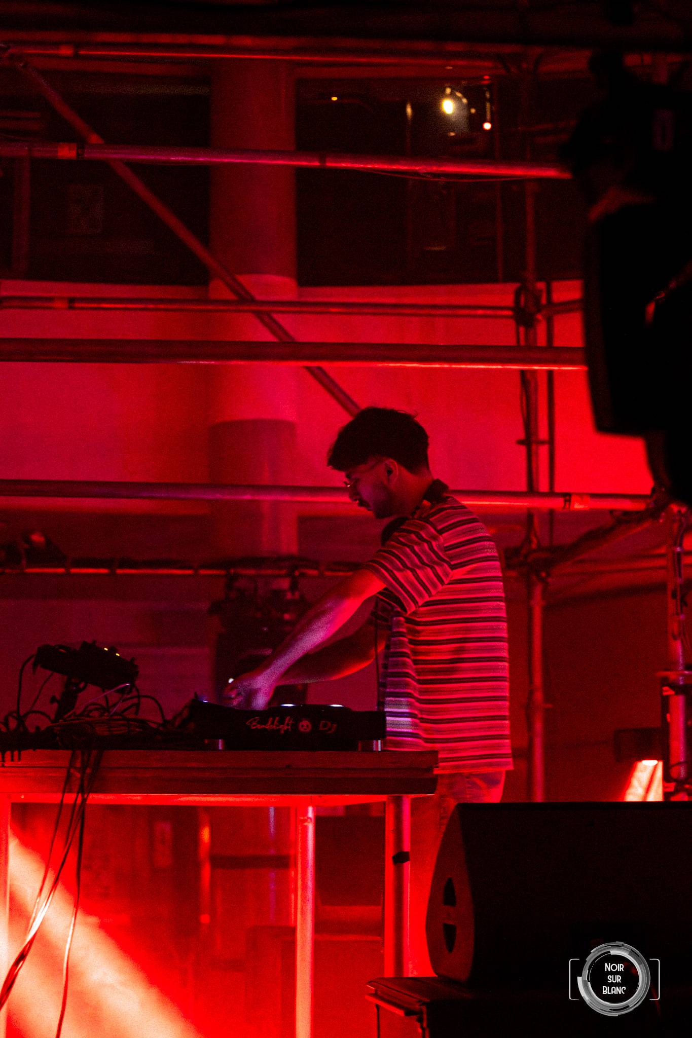 Le même DJ vu de profil, la salle entière noyée dans une lumière rouge, une enceinte de retour à ses pieds.
