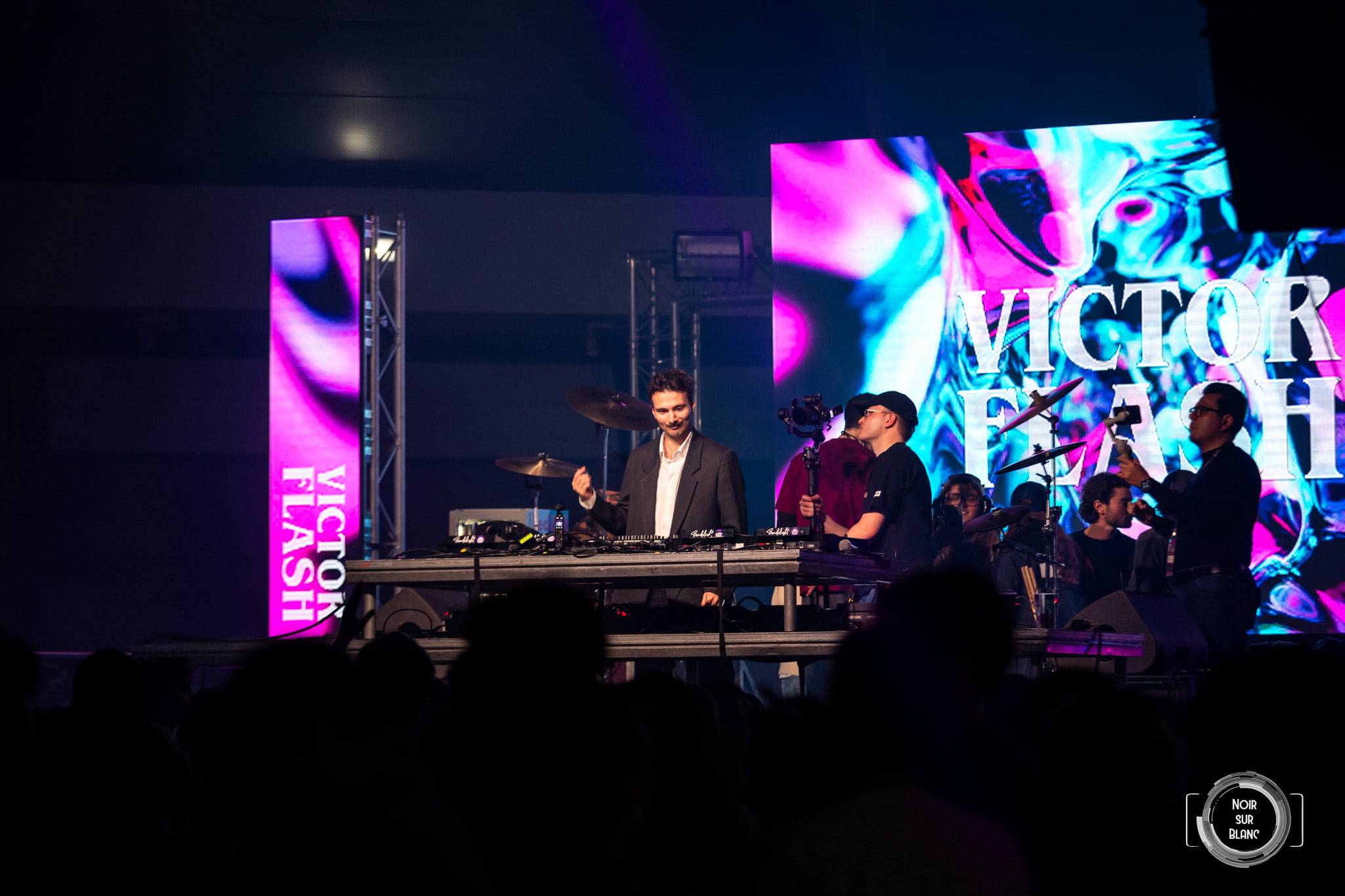 Sur scène, un DJ et un batteur jouent devant un écran géant affichant « Victor Flash » en lettres blanches.