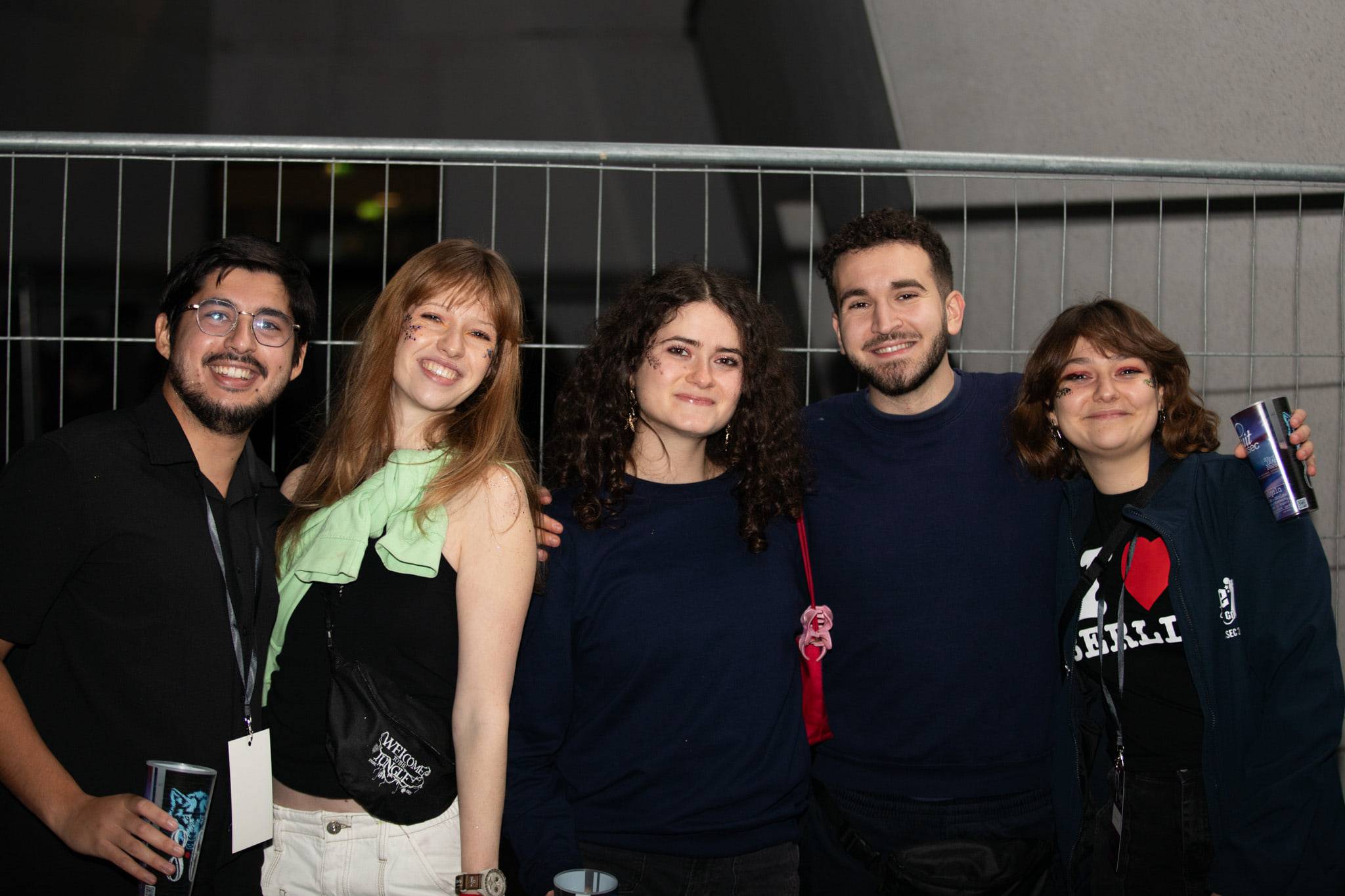 Cinq étudiants posent en souriant devant une grille métallique, gobelets à la main.