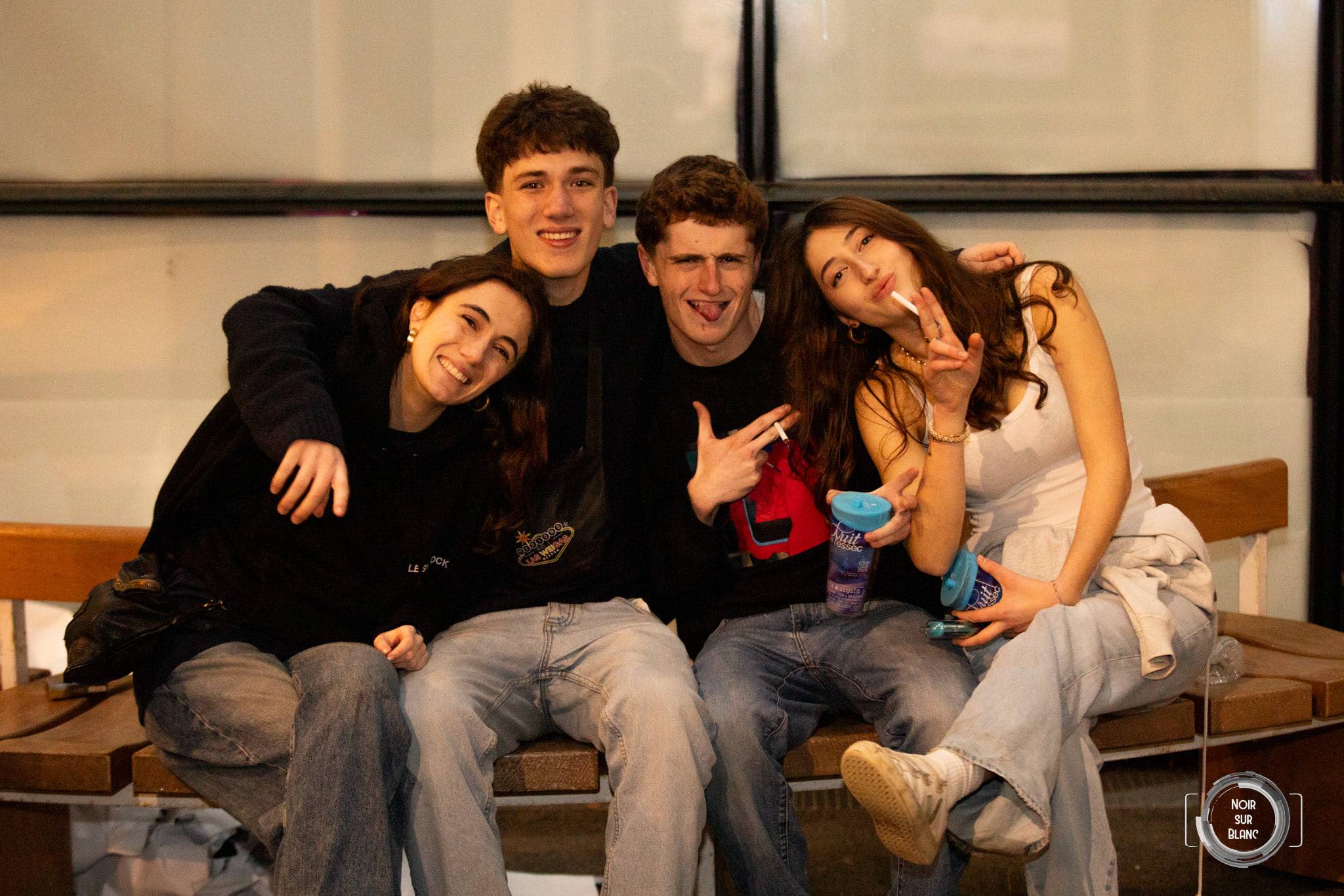 Quatre étudiants assis sur un banc de bois font des grimaces face à l'objectif, gobelets à la main.