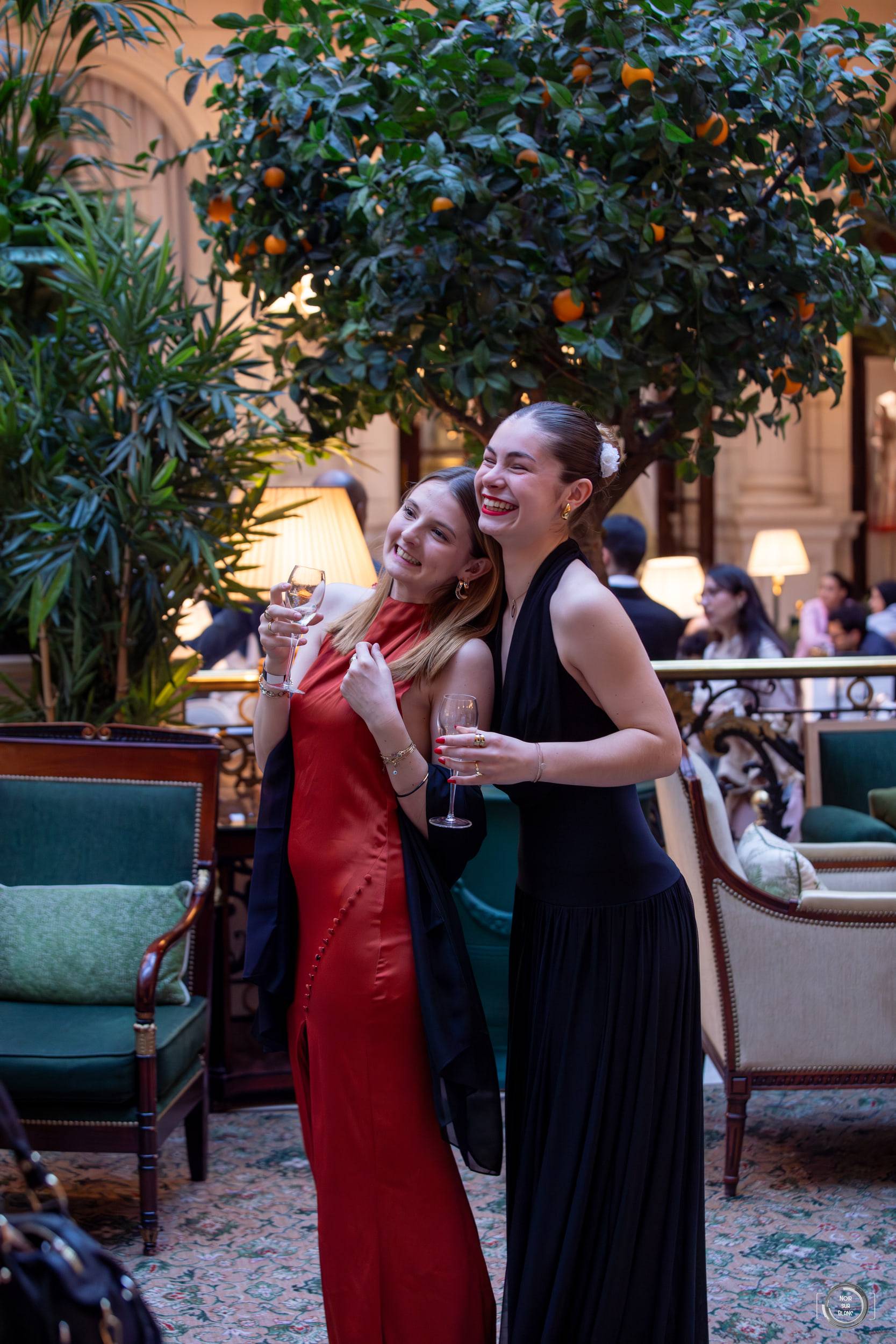 Deux étudiantes, l'une en robe rouge, l'autre en robe bleu nuit, posent enlacées devant un oranger en pot dans un salon d'hôtel.