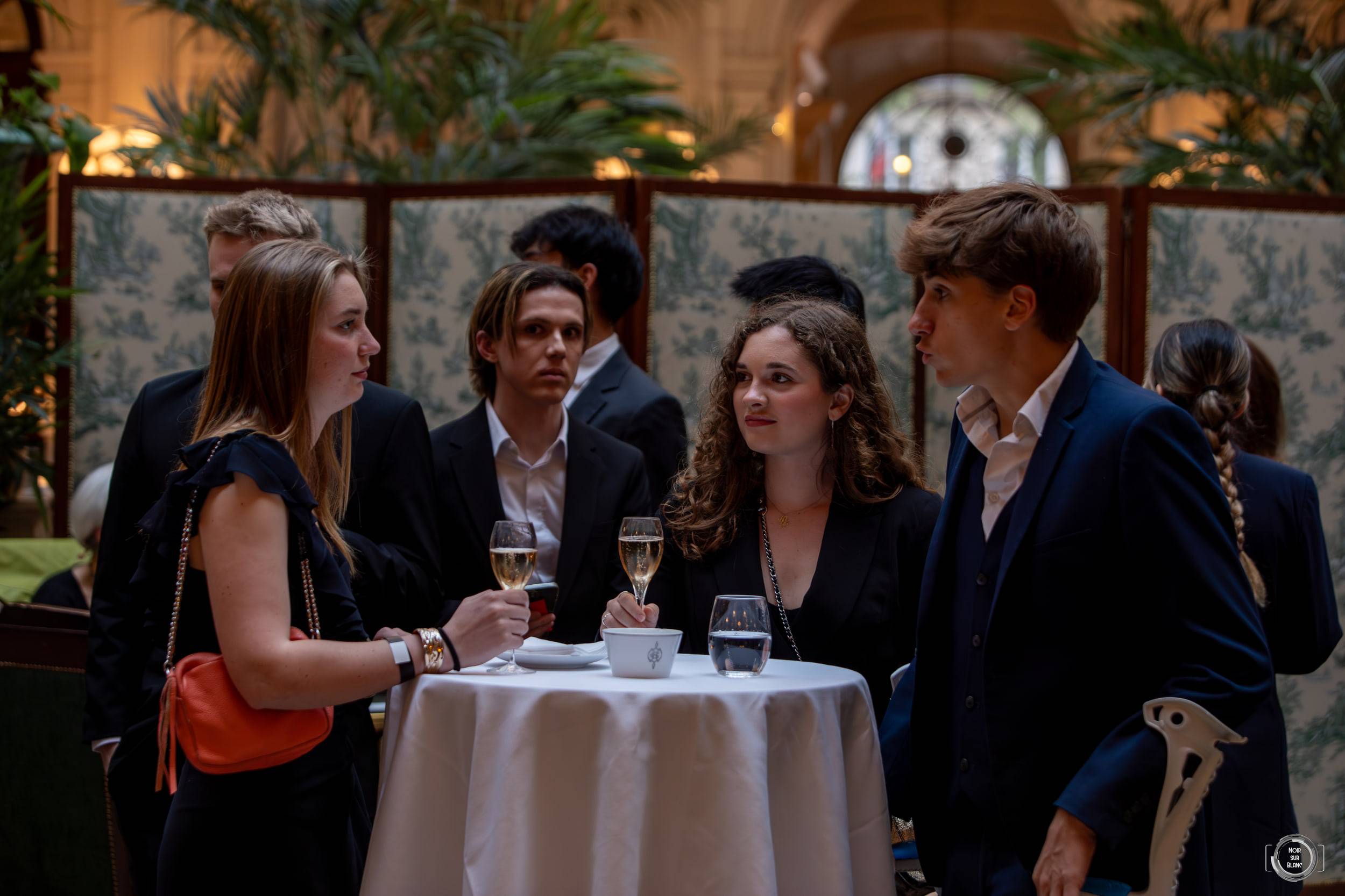 Quatre étudiants en costume et en robe de soirée discutent autour d'une table haute, une coupe à la main.