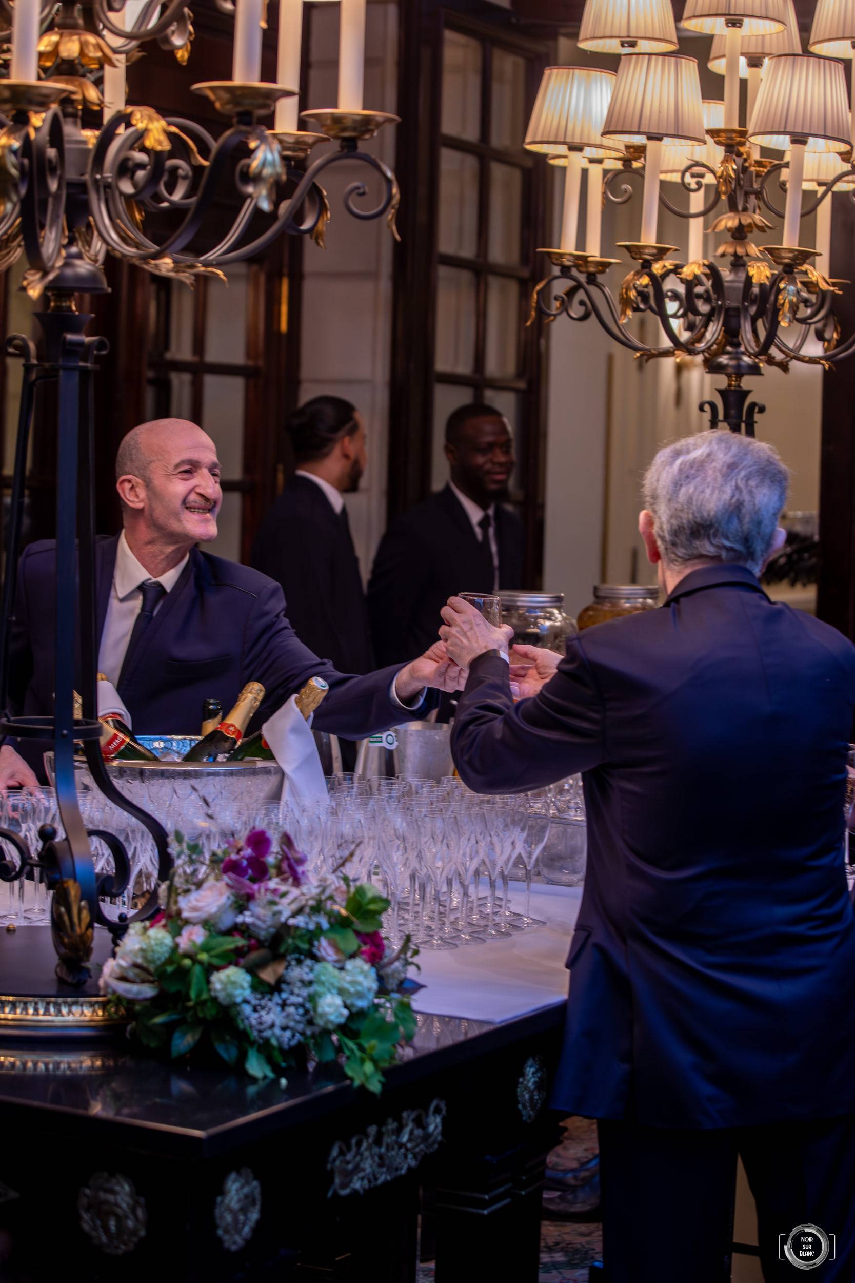Derrière un comptoir surmonté de candélabres et de bouquets, deux barmans préparent les verres de la soirée.