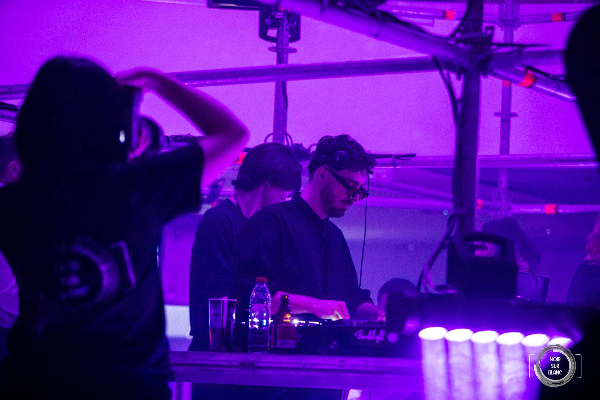 Deux DJ derrière les platines sous une bâche violette, une rangée de gobelets alignée devant eux.