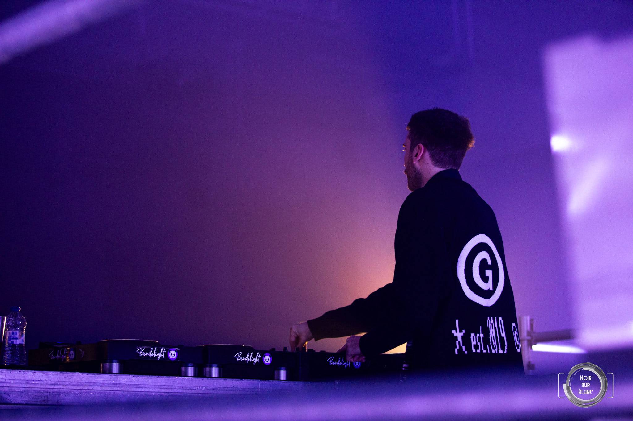 Un DJ de profil dans une lumière mauve, le dos de son blouson marqué d'un grand G.