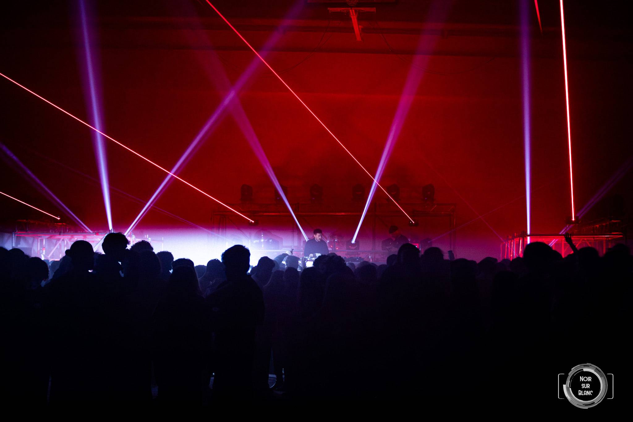 Vue d'ensemble de la salle noyée de rouge, traversée de lasers roses au-dessus des silhouettes du public.