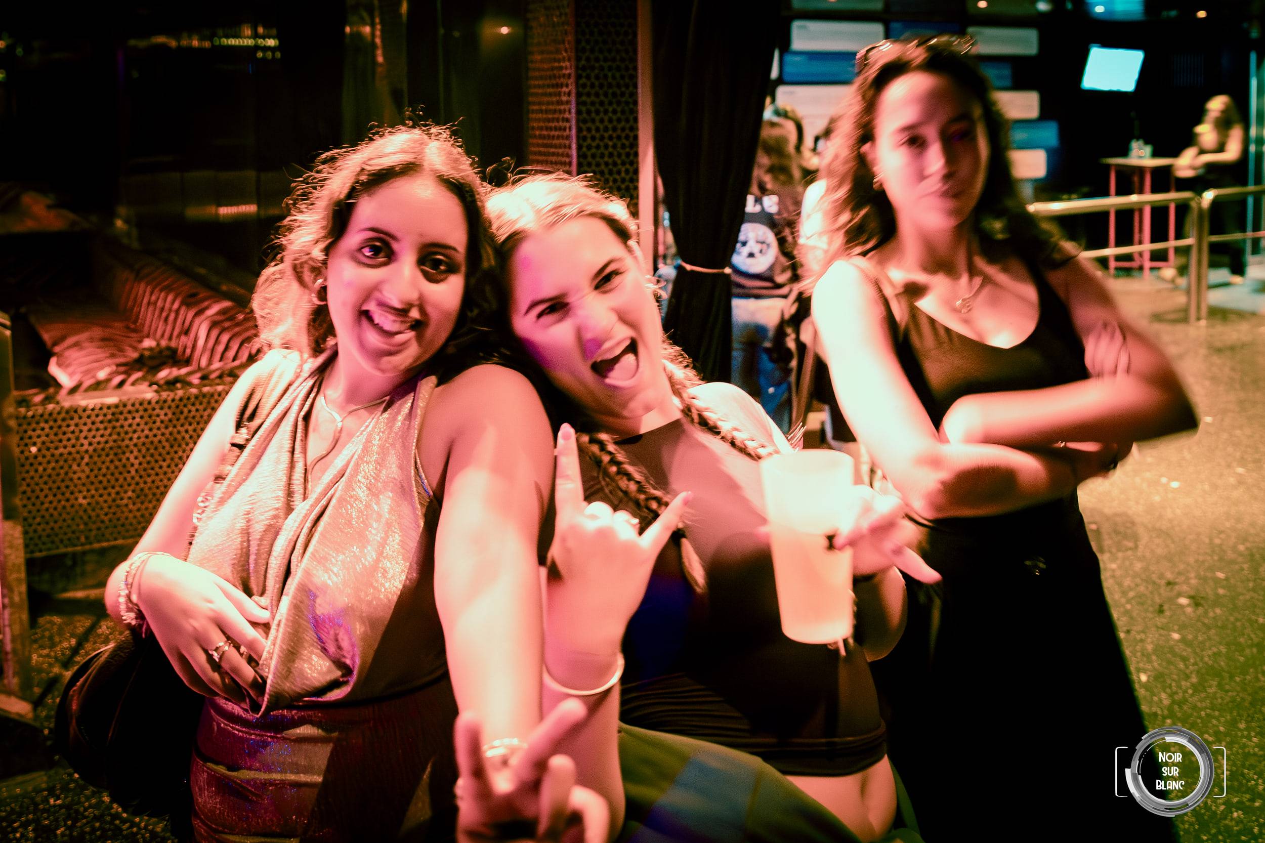 Trois jeunes femmes posent dehors dans la nuit, un gobelet à la main, devant une rambarde métallique.