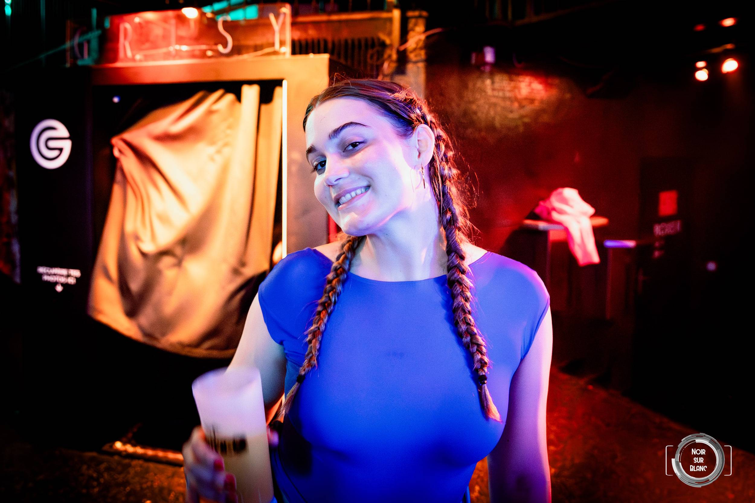 Une jeune femme aux longues tresses, en haut bleu, sourit face à l'objectif devant un mur éclairé de rouge.